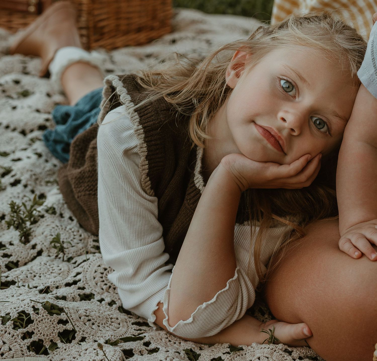 Blonde girl with blue eyes lying on a lace blanket at a picnic, resting chin on hand.