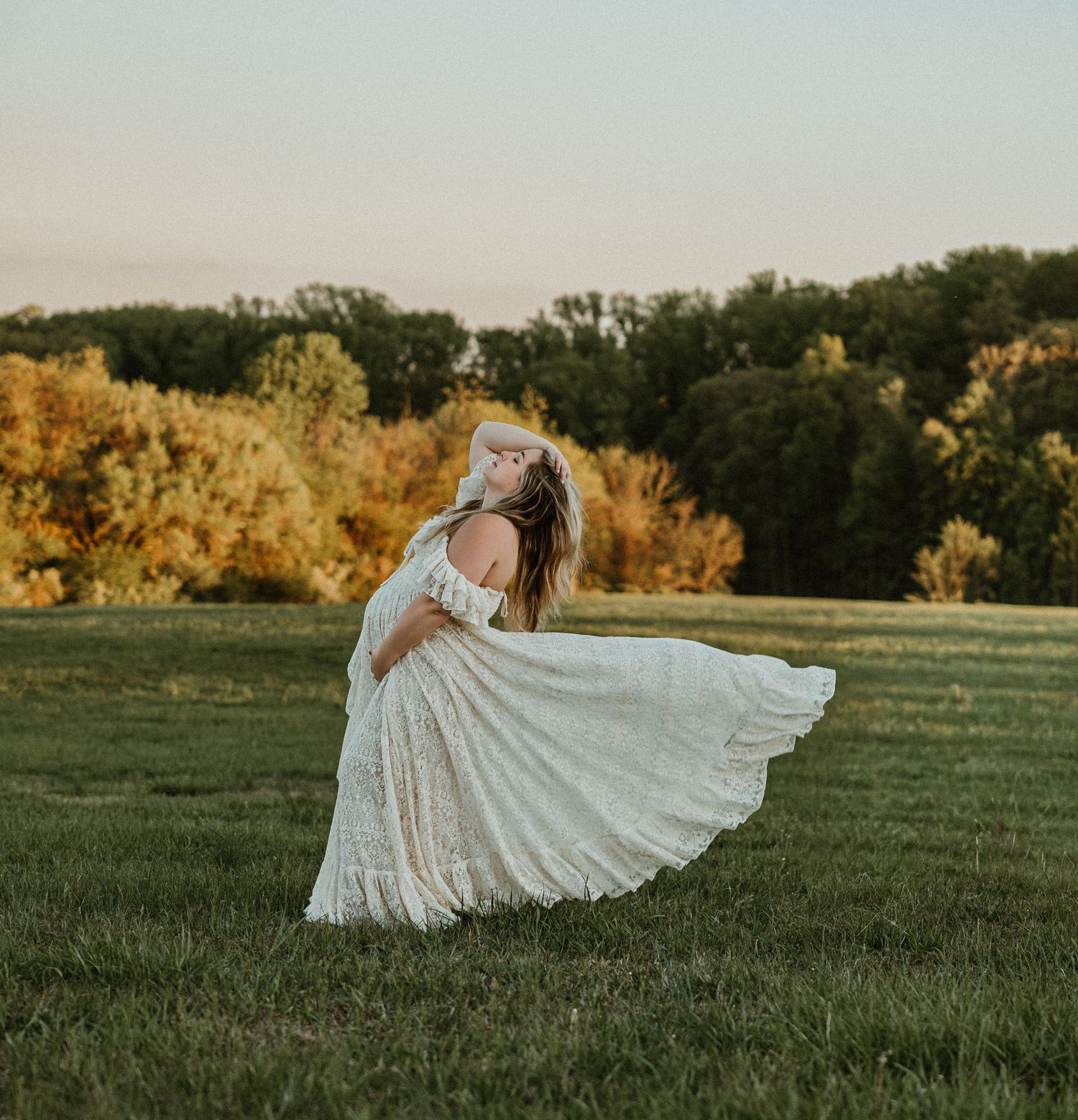 A bride in a flowing white lace gown twirls in a green meadow at golden hour with autumn trees in the background.