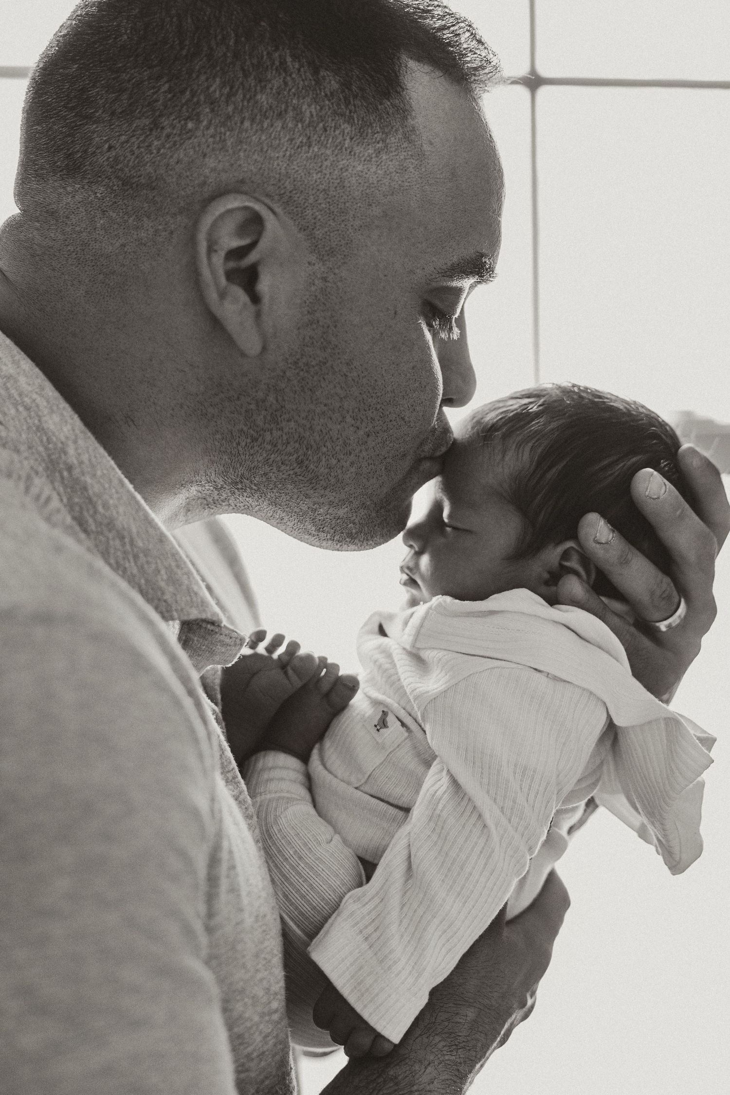 Black and white photo of a father tenderly kissing his newborn baby on the forehead.