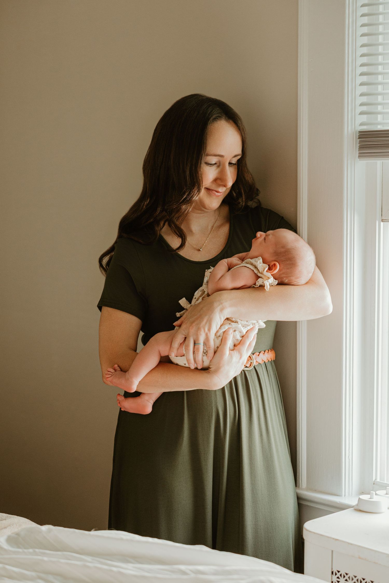 A smiling brunette mother in a green dress tenderly holds her newborn baby near a sunlit window.