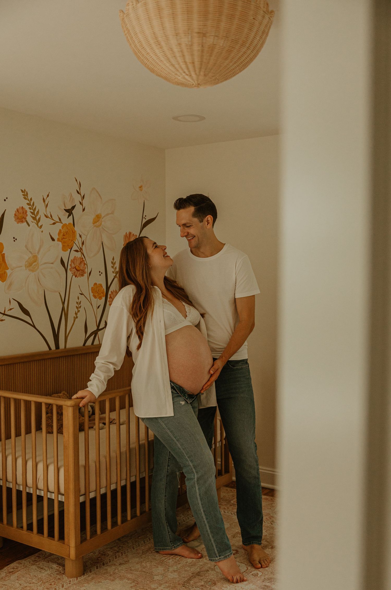 Expecting couple smiling in nursery with floral mural, wooden crib, and rattan pendant light.