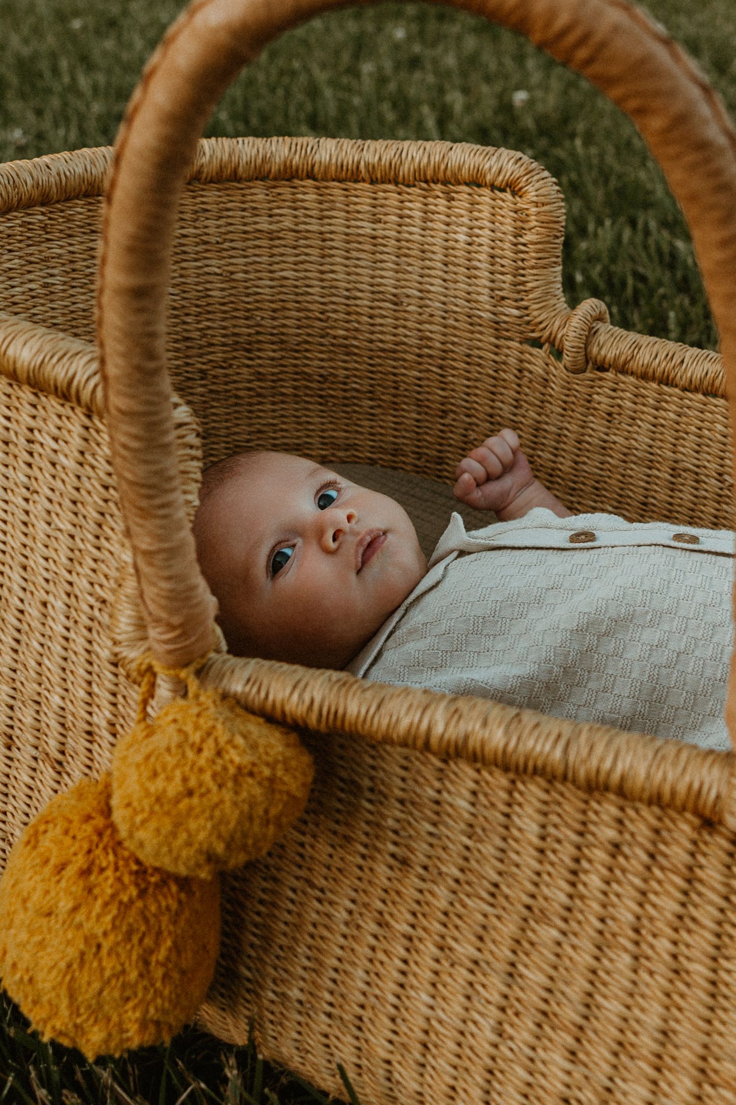 Baby lying in a wicker Moses basket with mustard yellow pom poms outdoors on green grass.