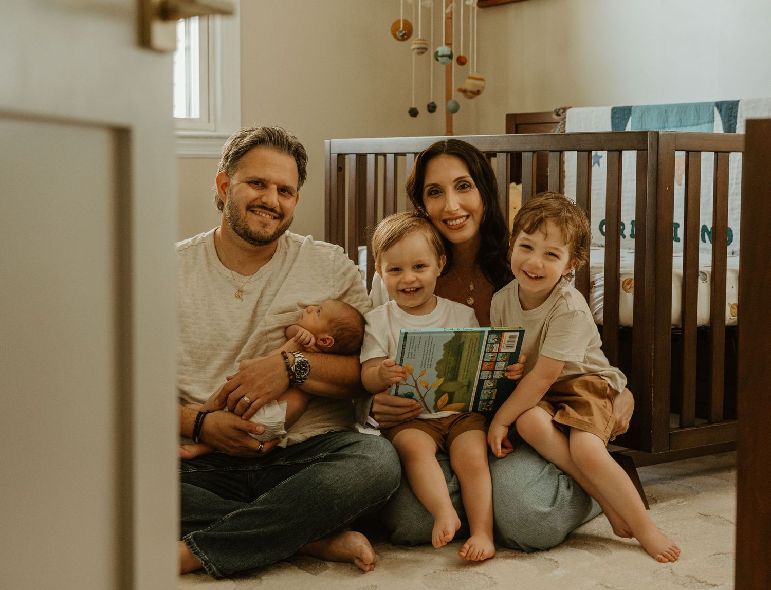 Happy family of five sits together in nursery, parents holding newborn while children smile.