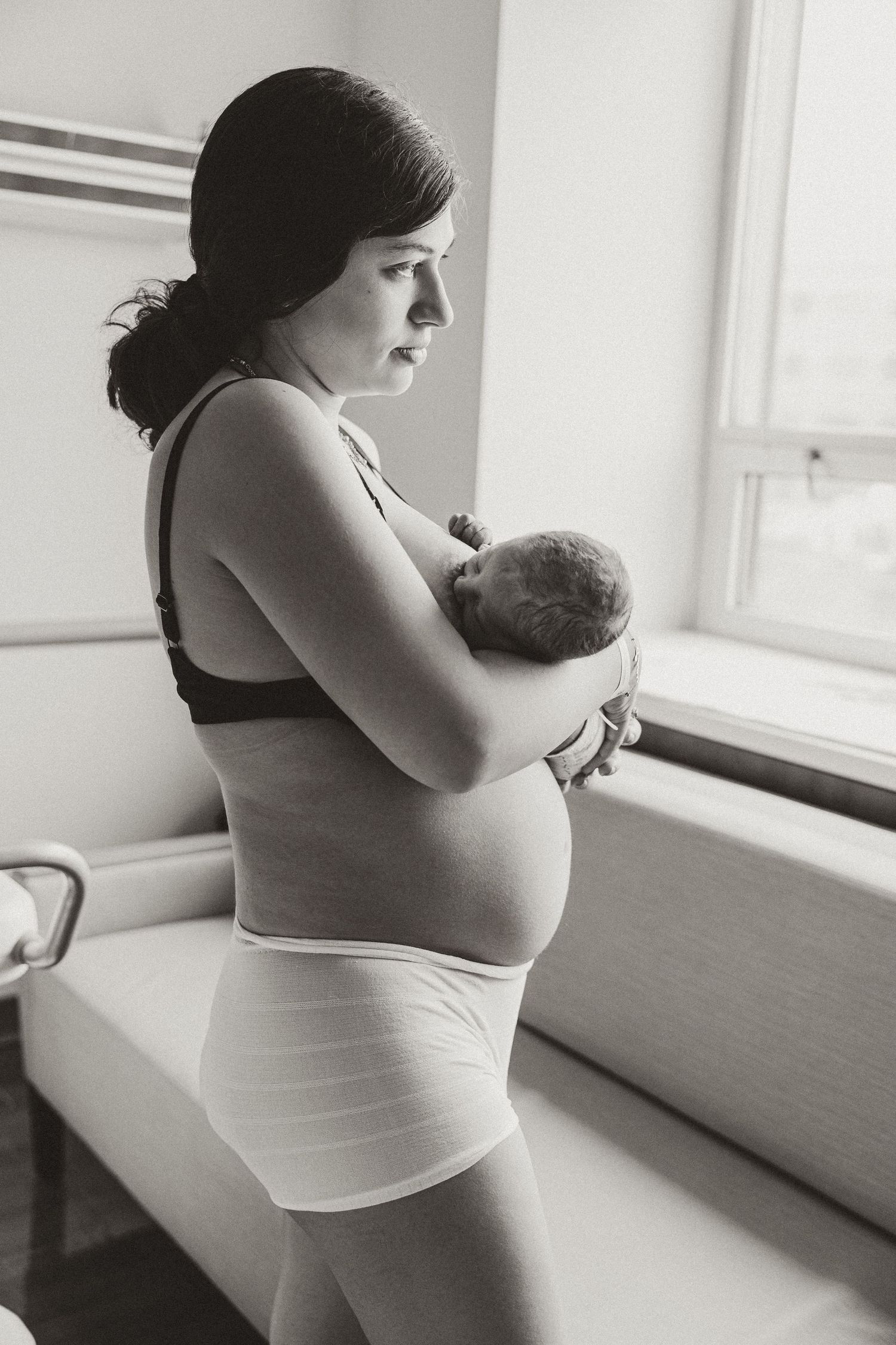 New mother holds her newborn baby while standing by a window in a hospital room postpartum.
