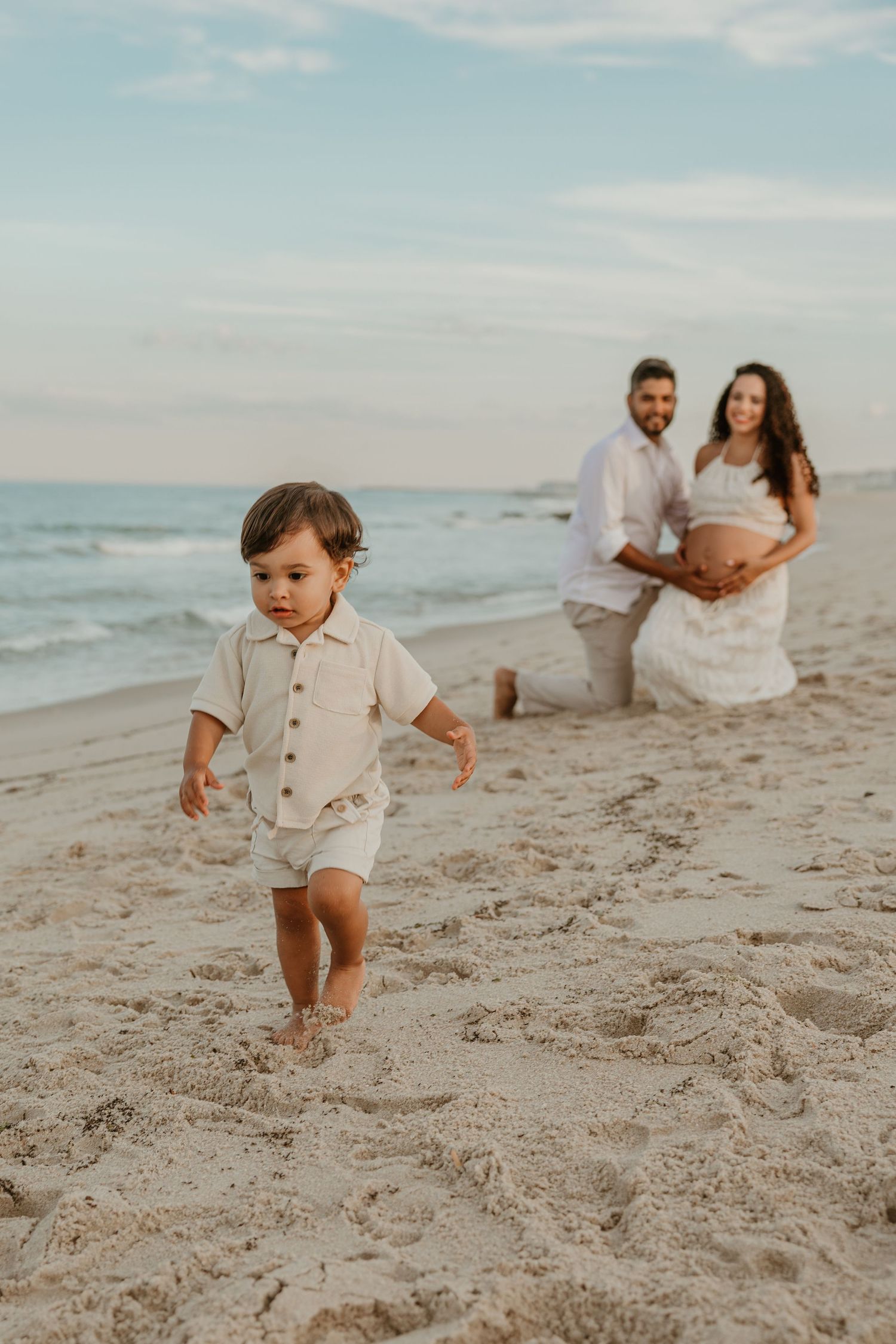 A toddler walks on a sandy beach while his parents, including a pregnant mother, kneel together in the background.