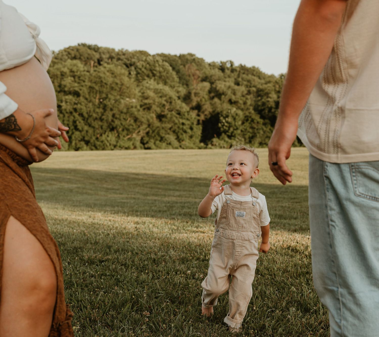 A happy toddler in overalls runs toward pregnant mother and partner in a sunny green field.