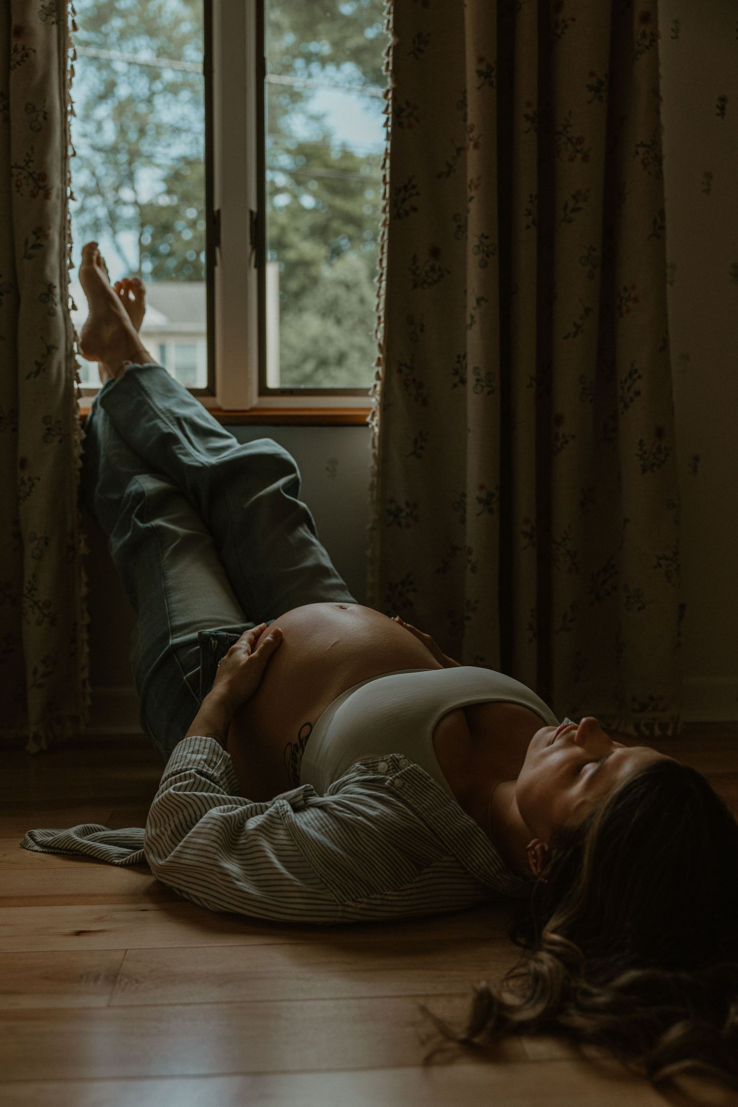 Pregnant woman lying on hardwood floor near window, cradling baby bump in soft natural light with floral curtains.