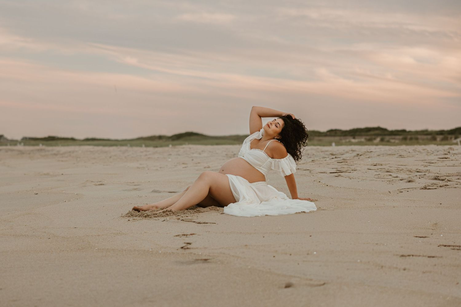 Pregnant woman in white dress sitting on sandy beach at sunset, posing gracefully with hand in hair.