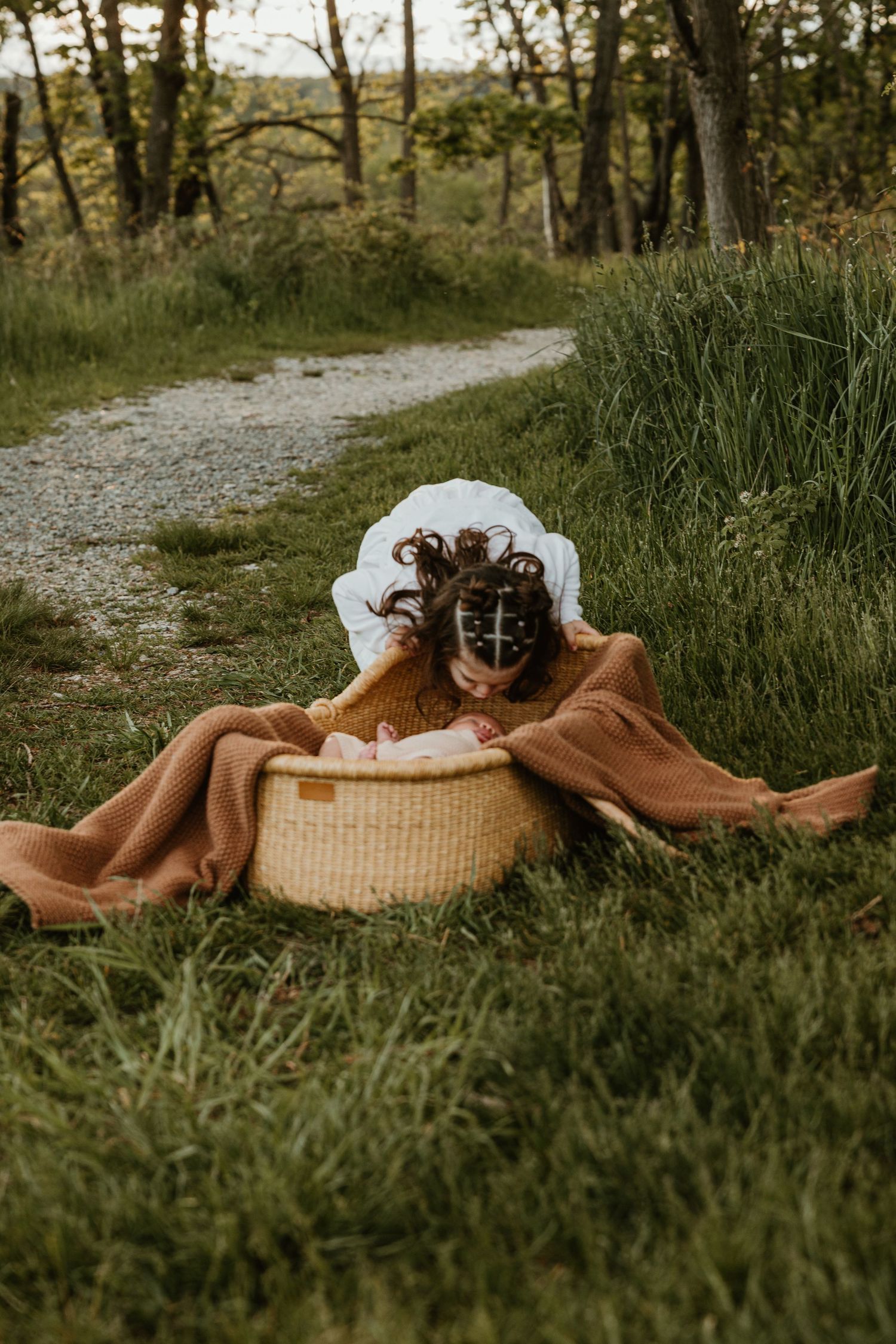 Young girl in rust dress and white bonnet leaning over wicker basket on grass in nature.