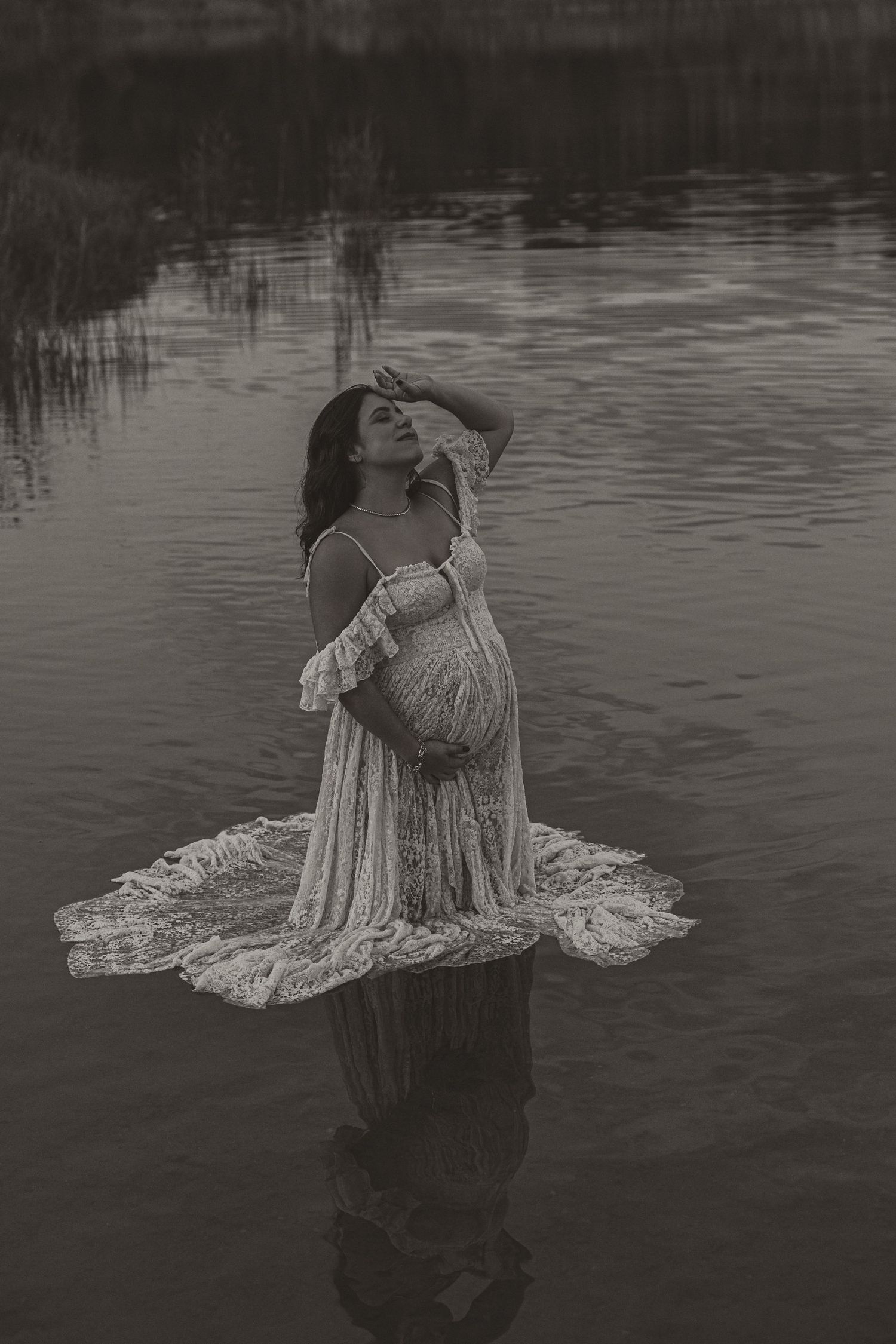 Pregnant woman in lace gown standing in water, black and white maternity photo.