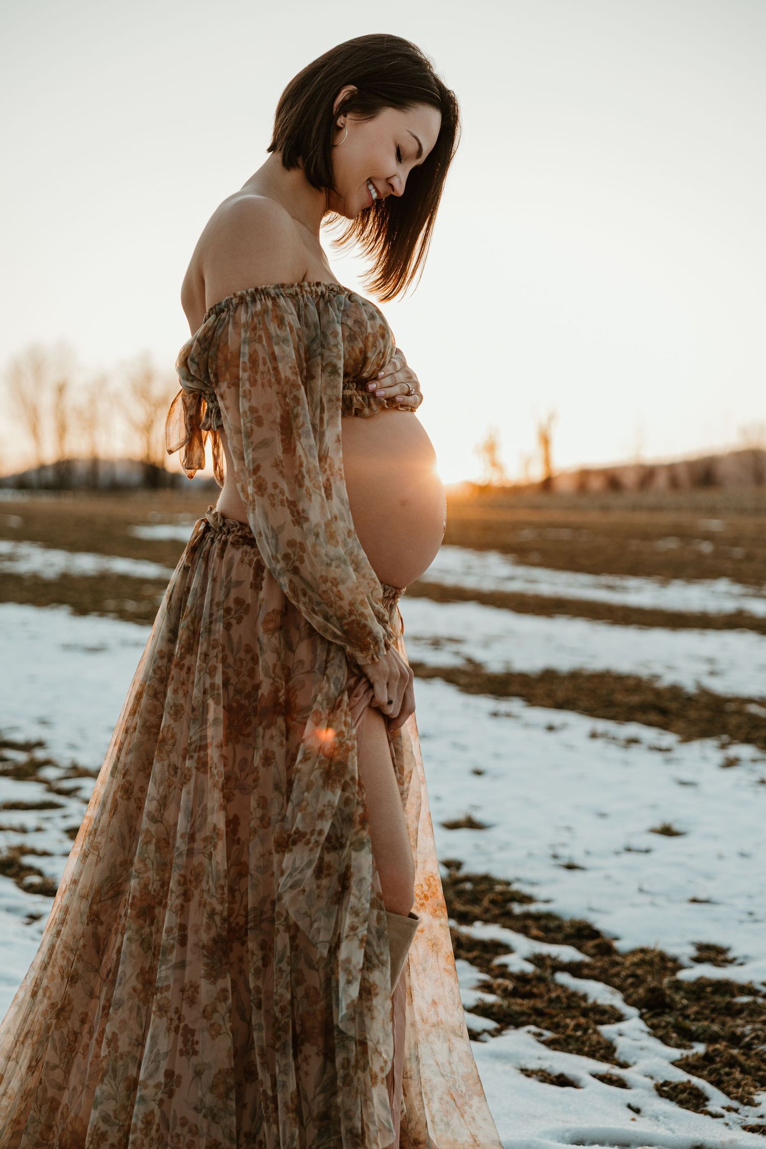 A person in a flowy floral dress stands in a snowy field at sunset, creating a dreamy winter portrait.