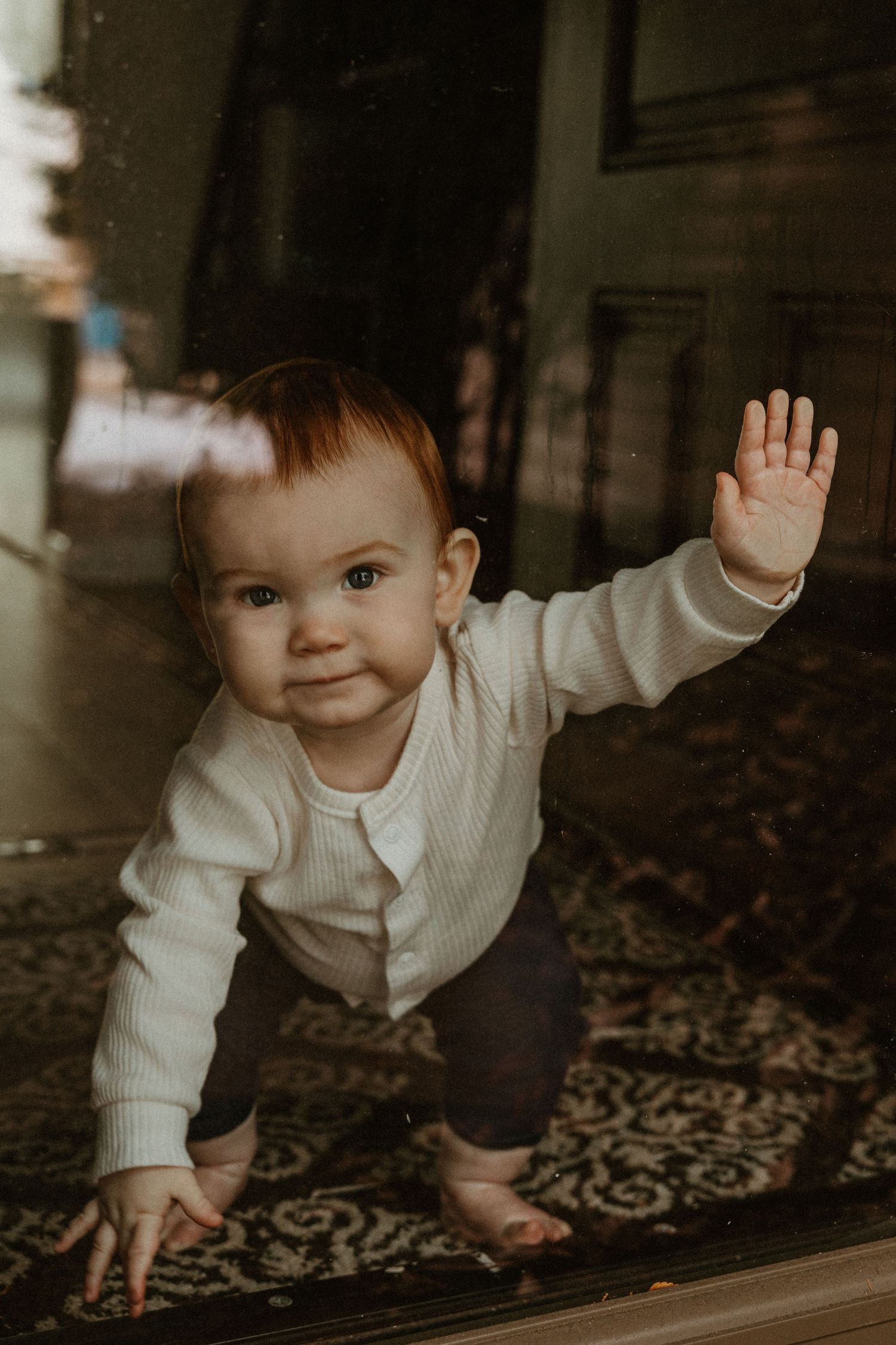 Baby in white sweater crawls and waves on patterned carpet.