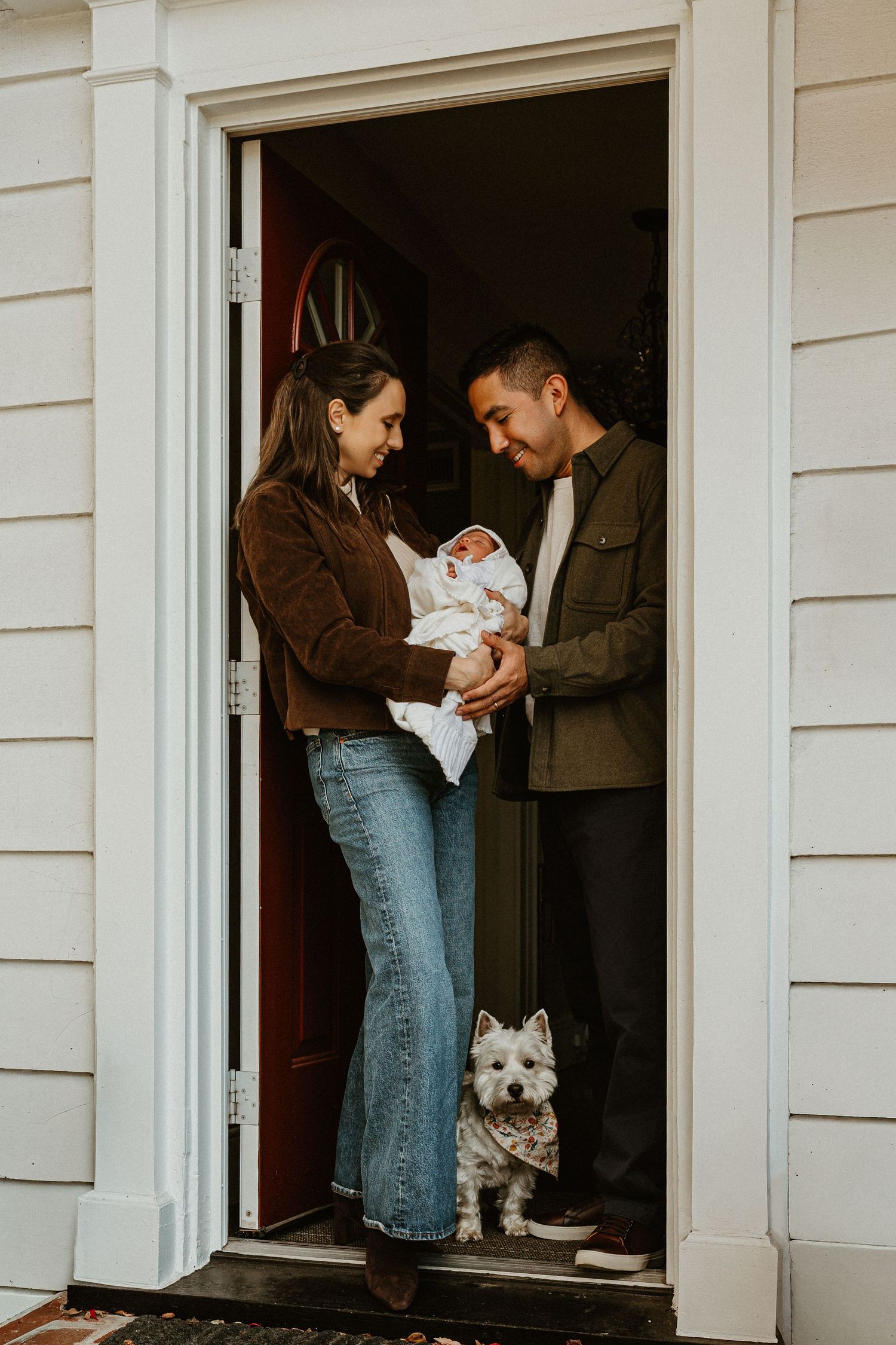 Family and dog welcome a new baby at their front door with white siding.