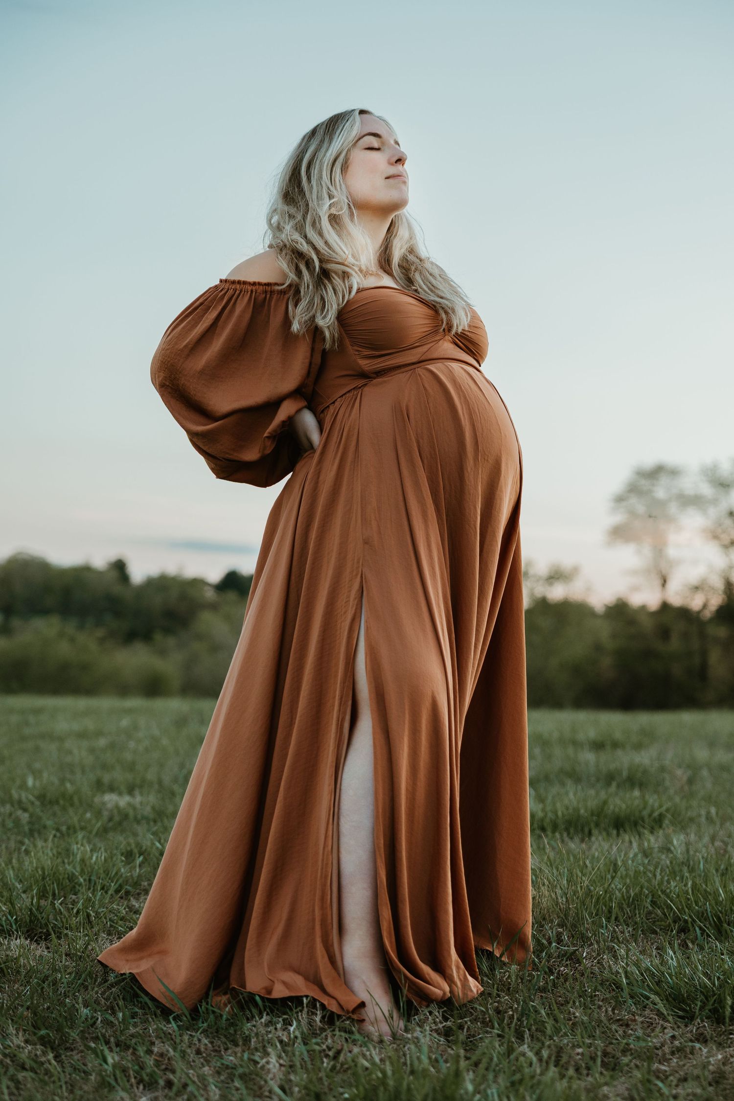 Pregnant woman in rust orange off-shoulder gown poses gracefully in a green field at dusk.