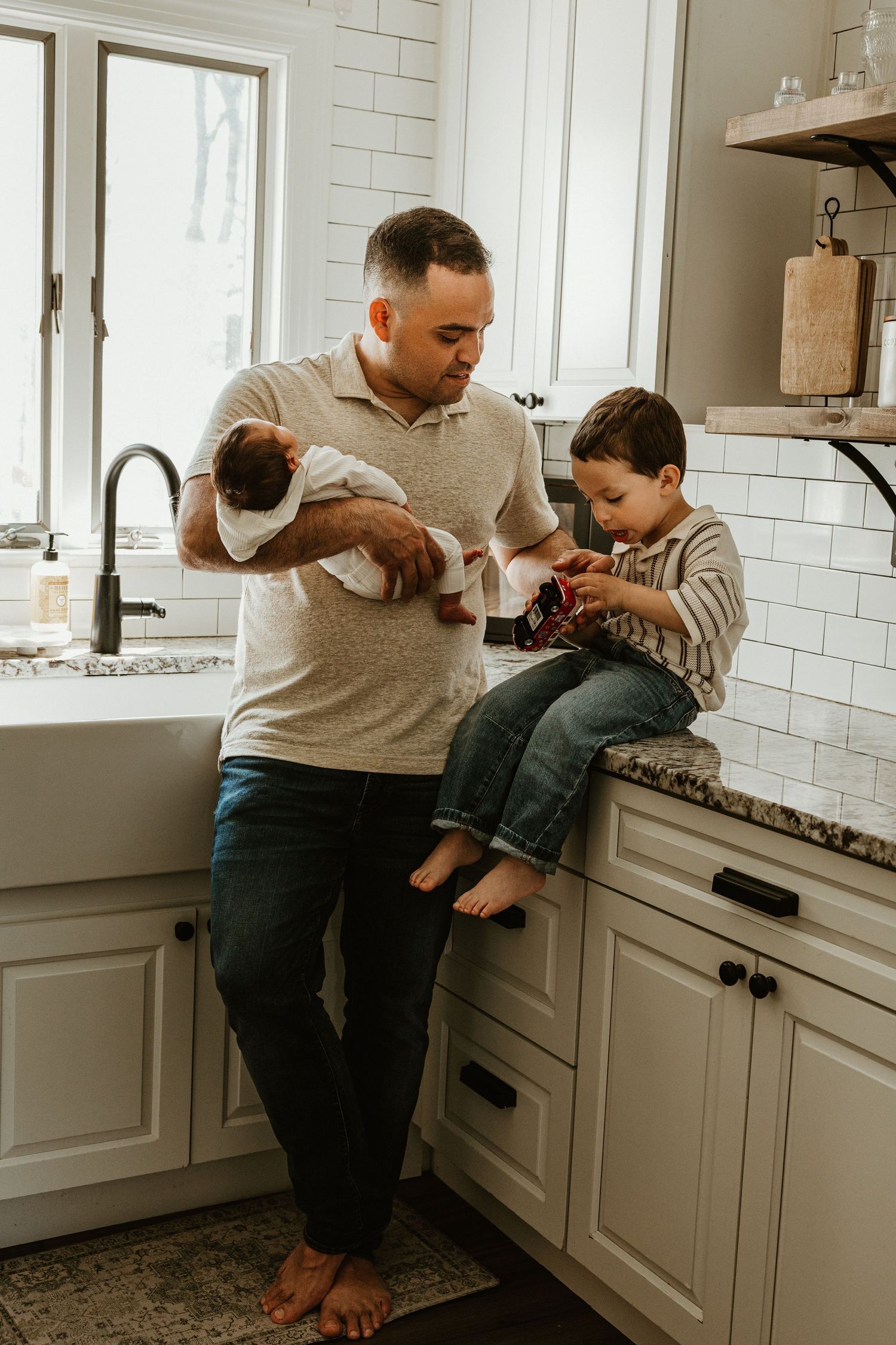 Father holds newborn baby while toddler son sits on kitchen counter in bright white farmhouse kitchen.