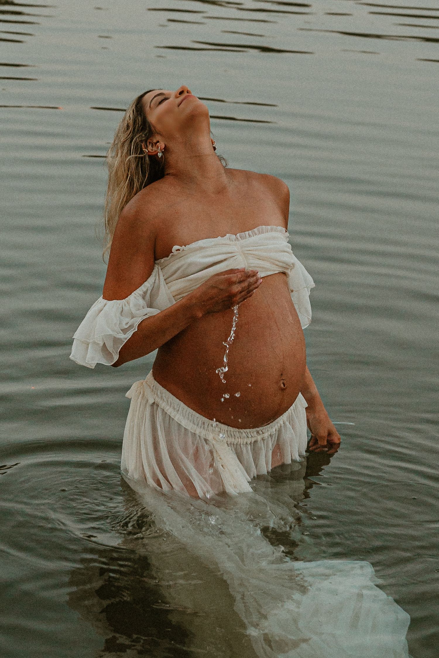 Pregnant woman in white off-shoulder dress standing in calm water, tilting head back with water dripping.