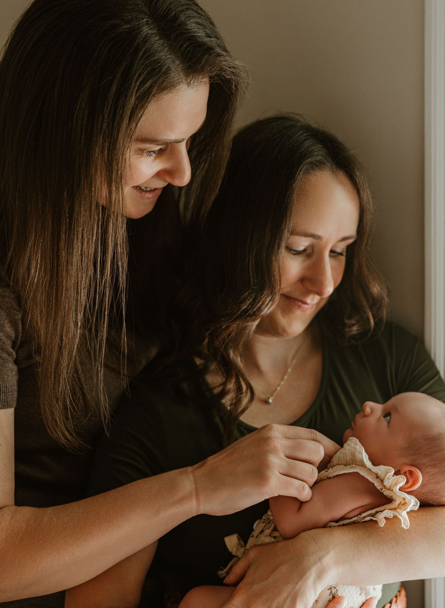 Two women in black tops lovingly gaze at a newborn baby held in their arms indoors.