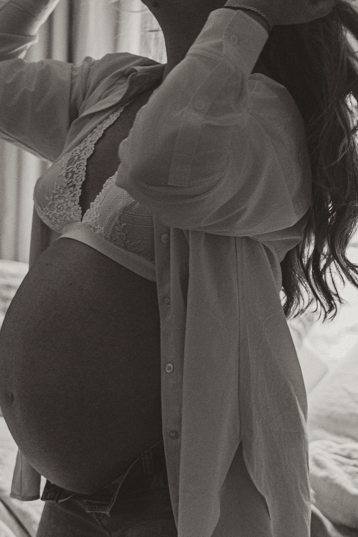 Black and white maternity photo of pregnant woman showing baby bump, wearing lace bralette and open button-up shirt.