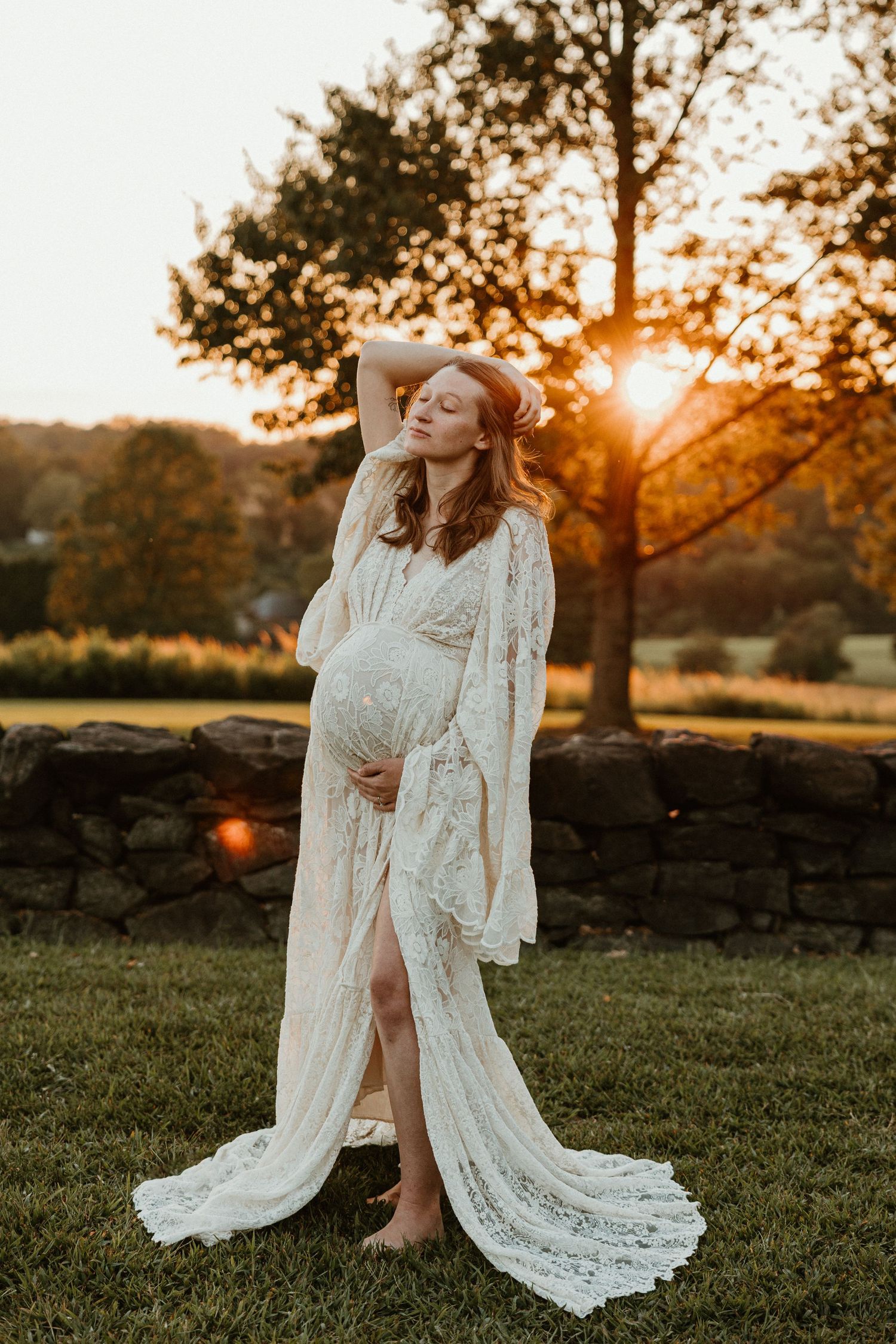 Maternity photo of expectant mother in white lace dress posing at sunset in a garden setting.