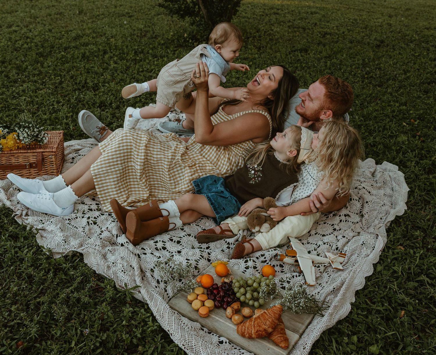Family laughing on lace blanket at outdoor picnic with fruits and basket on green grass.
