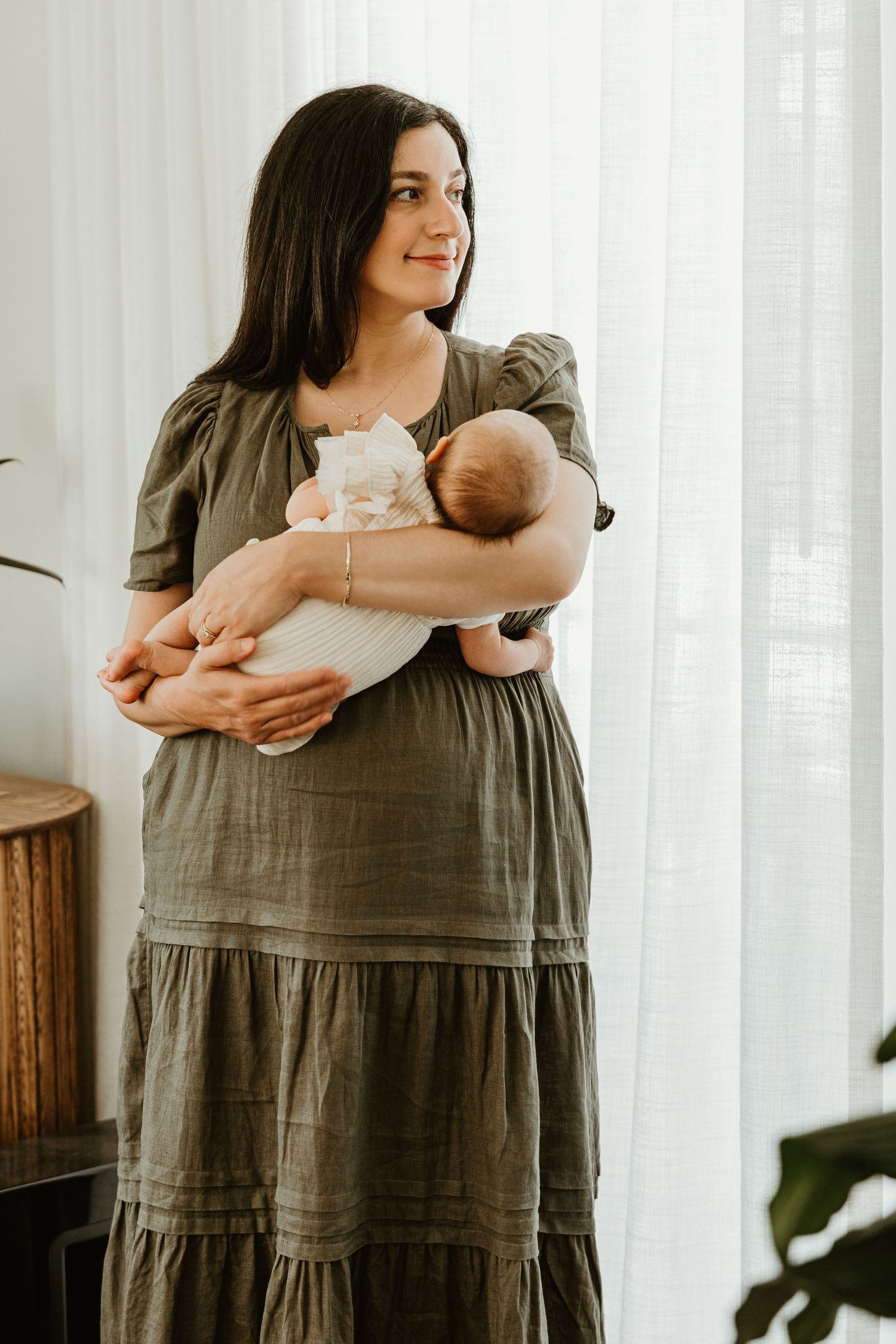 A smiling brunette mother in an olive green dress tenderly holds her newborn baby near a bright window.