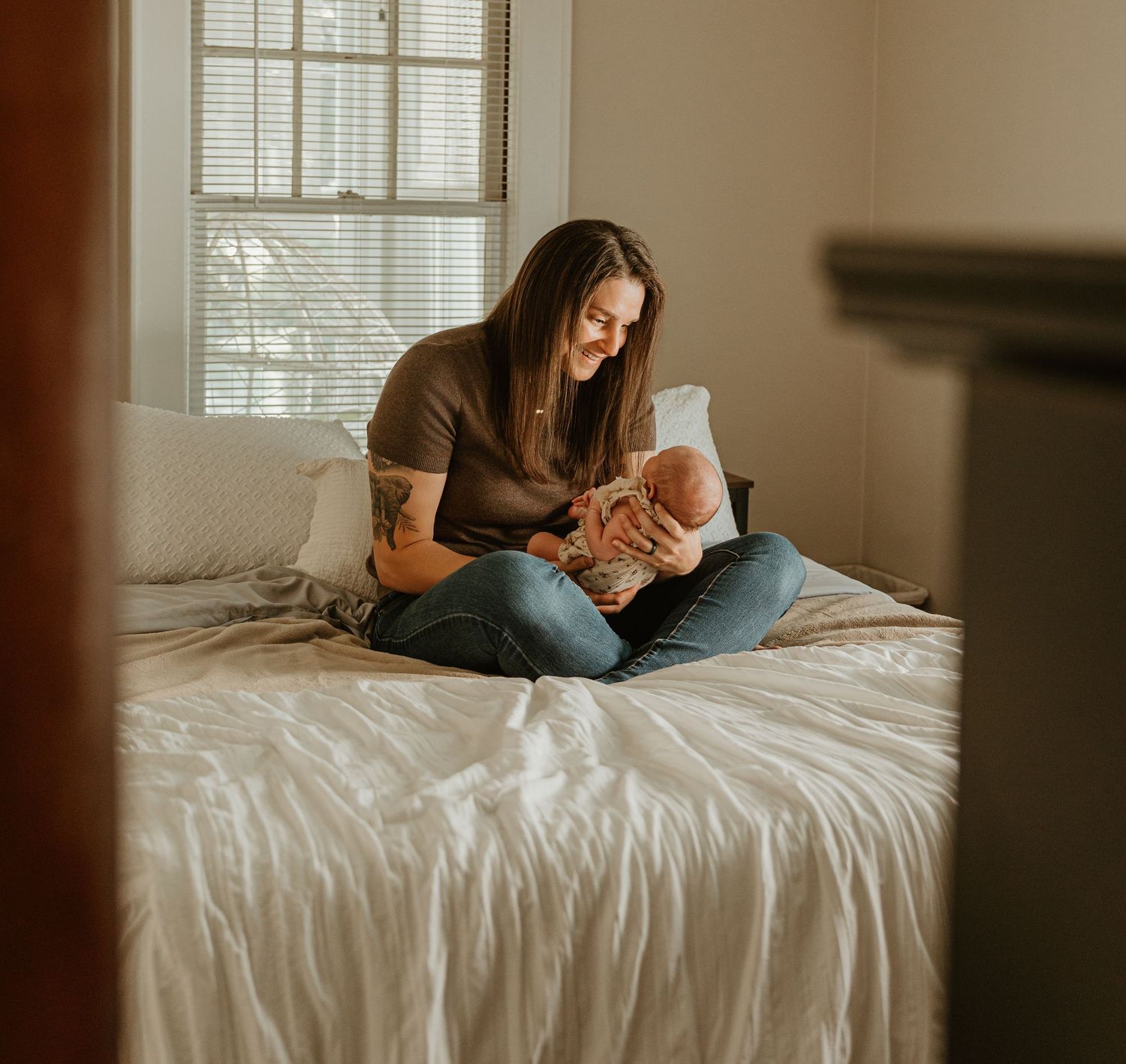A mother sits cross-legged on a white bed, smiling while cradling her newborn baby near a bright window.