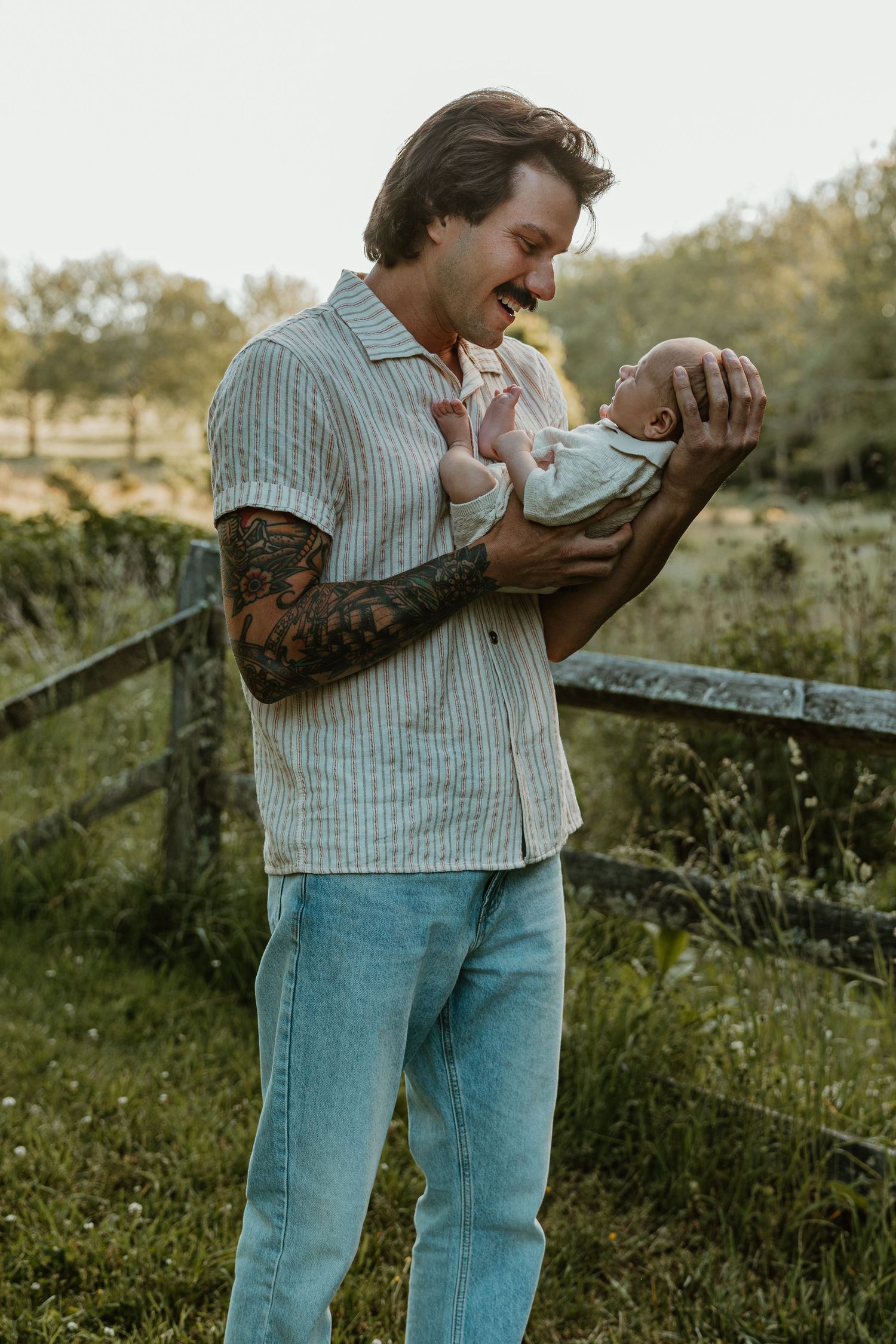 Tattooed man in striped shirt and jeans smiling while holding a newborn baby outdoors near a wooden fence.