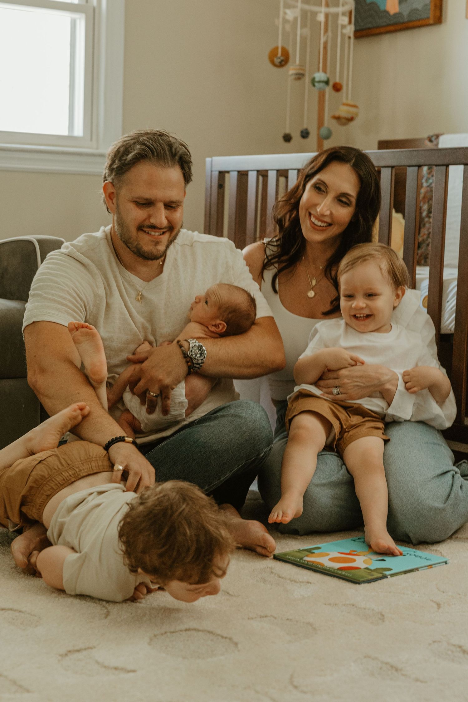 Happy family with newborn baby and toddlers sitting on nursery floor near crib, sharing a joyful moment together.