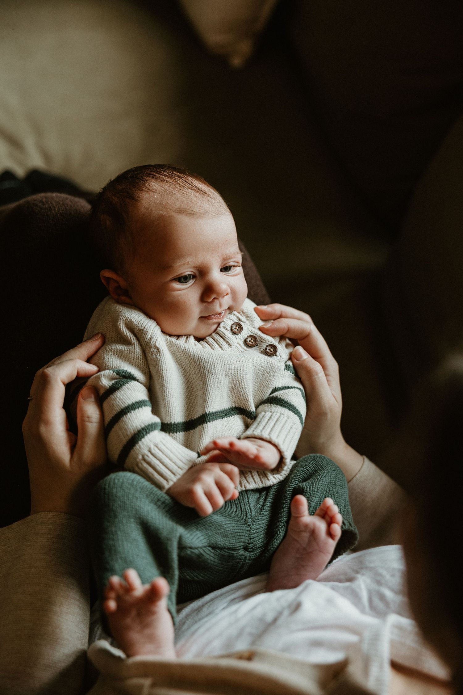 Baby in white knit sweater and sage green pants lying on soft bedding in dim light.