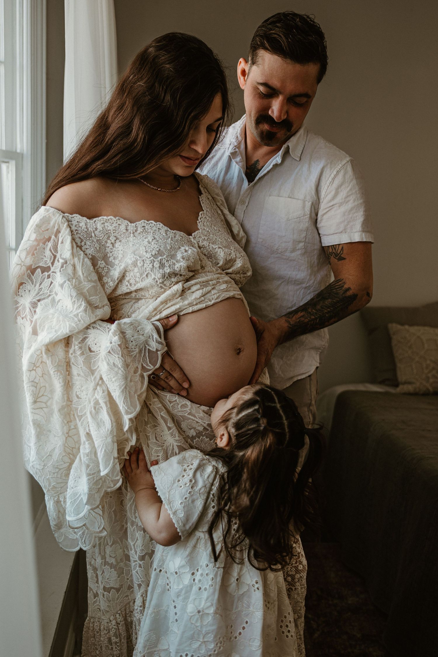 Intimate maternity session featuring white lace dress and soft window lighting.