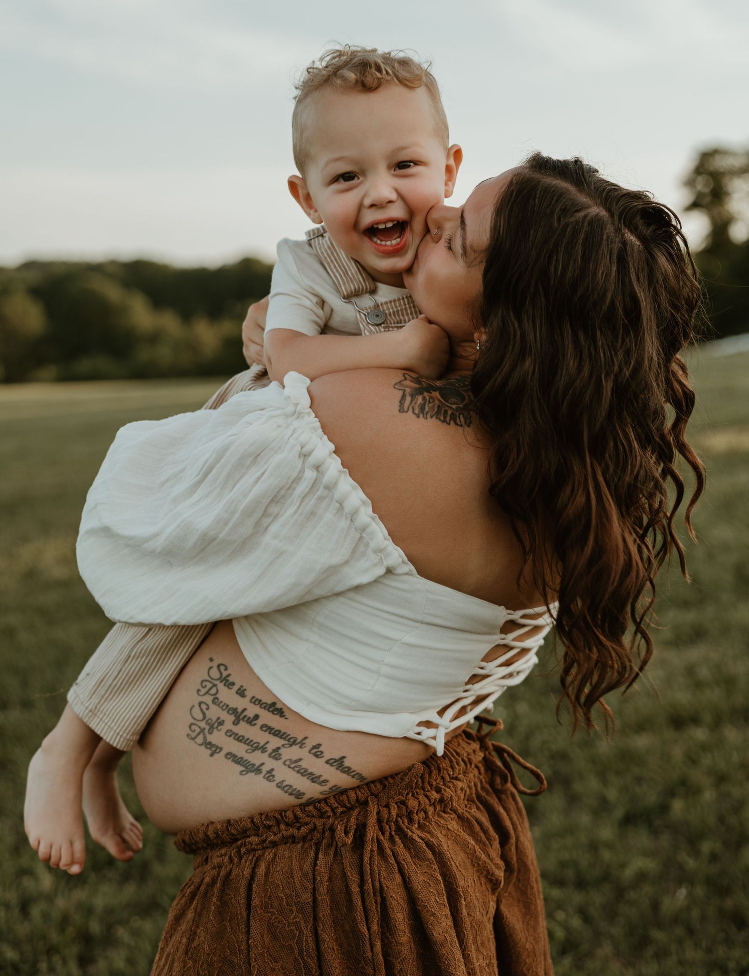 A joyful mother kisses her laughing baby boy outdoors, wearing a white off-shoulder top with a script tattoo.