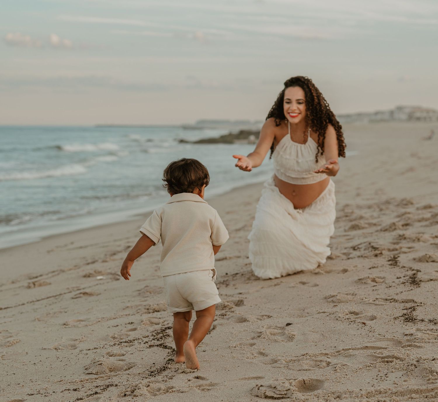 Pregnant woman in white dress crouches on sandy beach, reaching out to toddler running toward her at sunset.