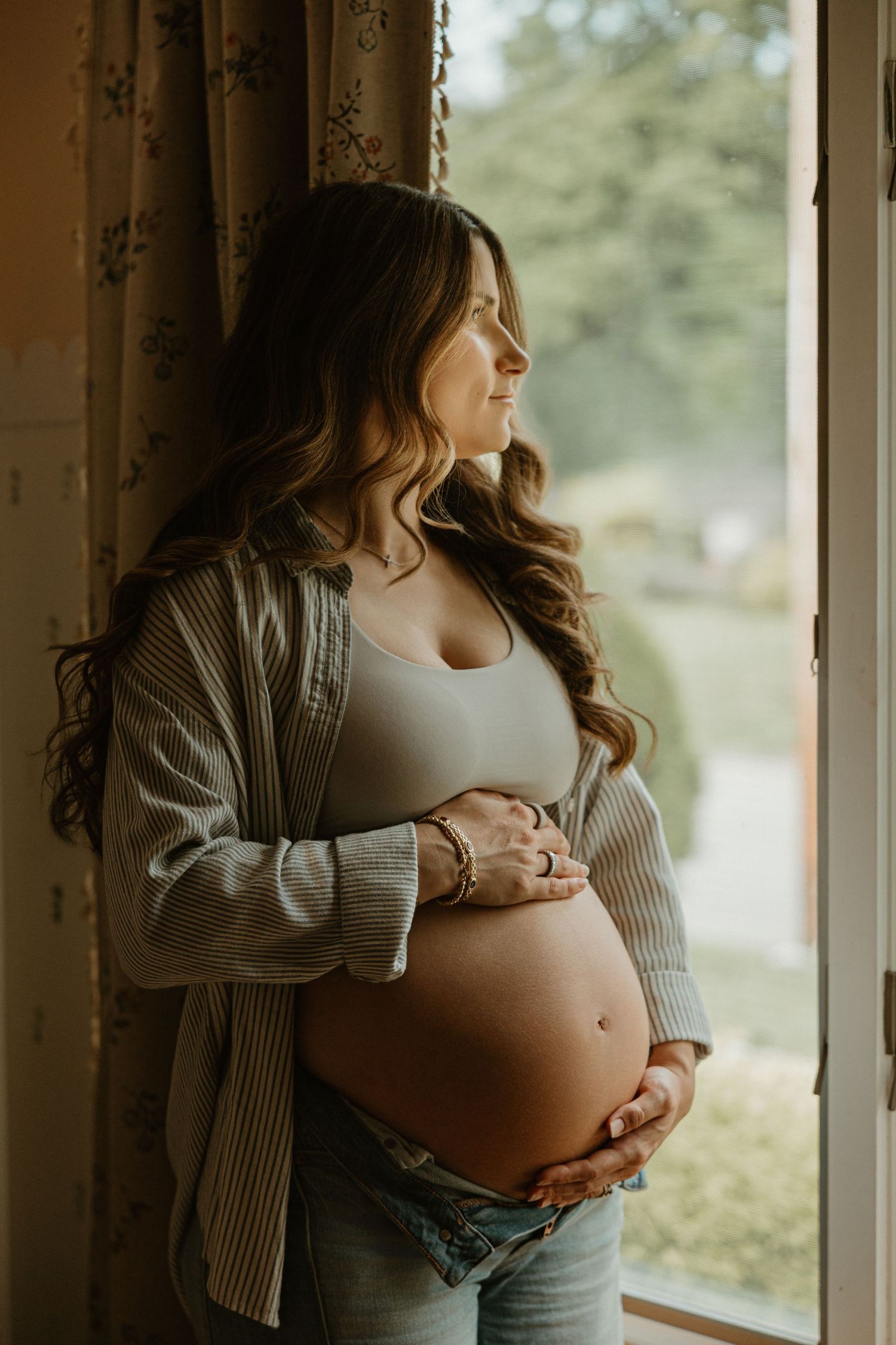 Pregnant woman in gray cardigan and jeans cradling her baby bump while gazing out a sunlit window.