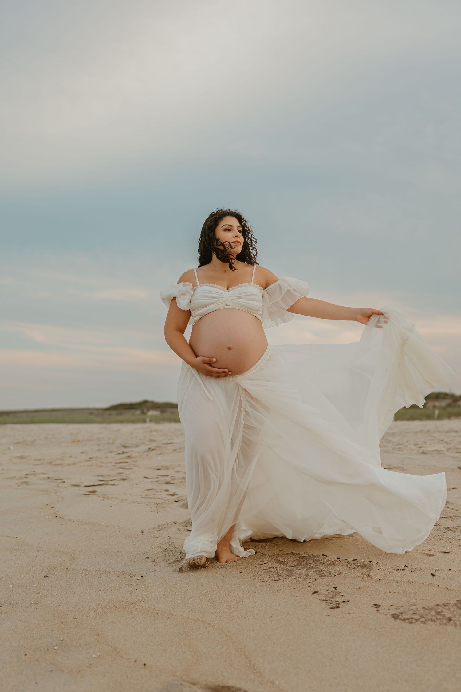 Pregnant woman in flowing white off-shoulder gown poses on sandy beach, cradling belly as dress billows in ocean breeze.