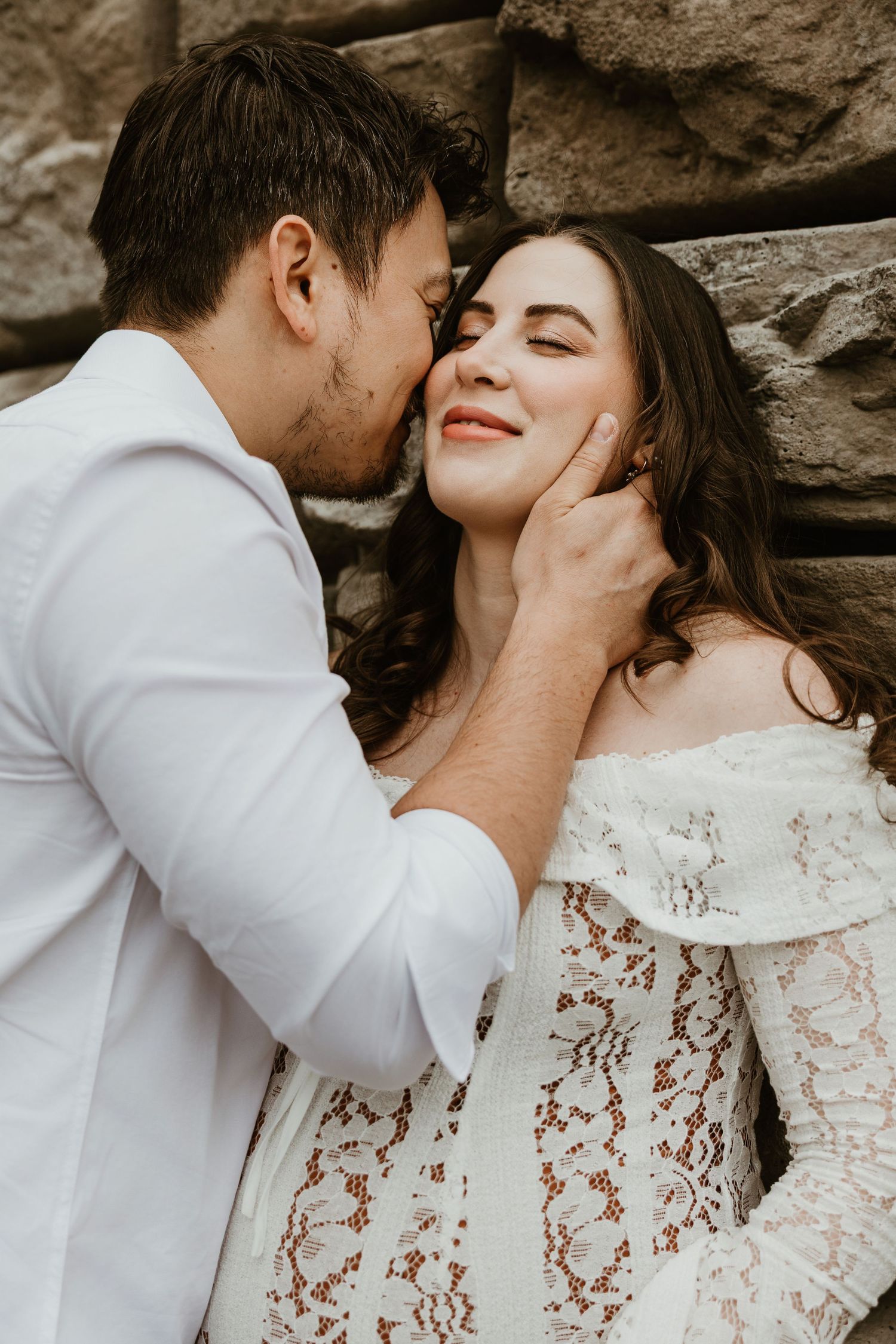 A groom tenderly kisses his bride on the cheek as she smiles, both dressed in white near a stone wall.