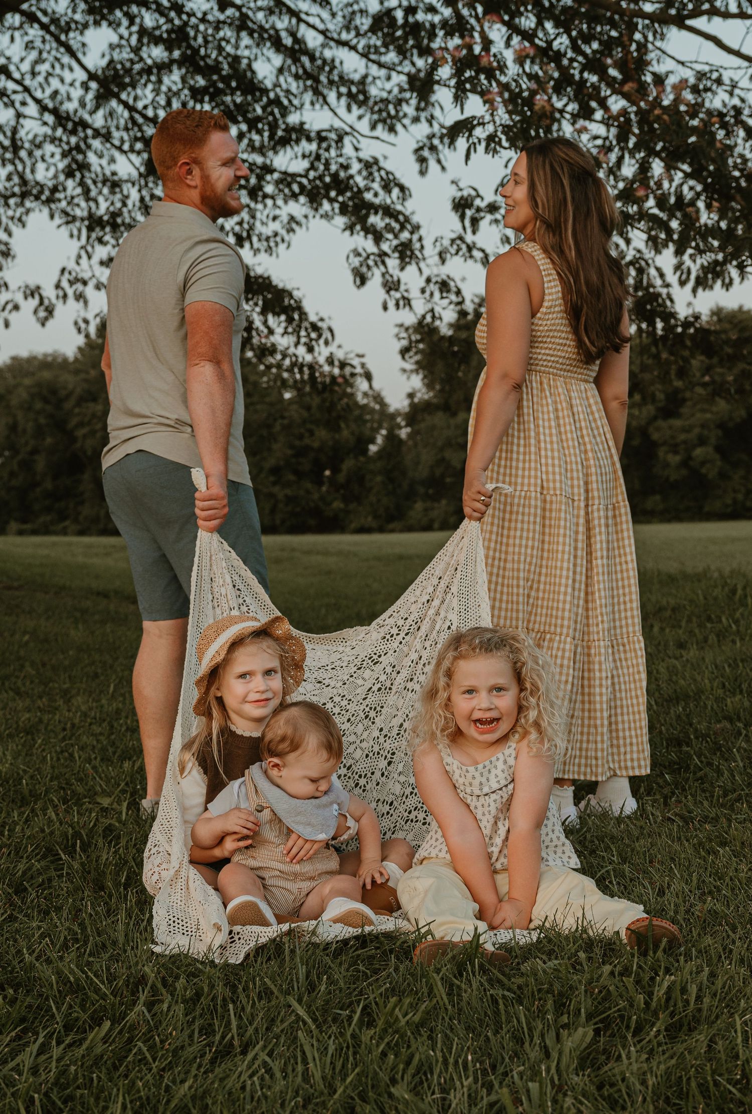 Family of five outdoors at sunset, parents holding lace blanket over three young children on grass.