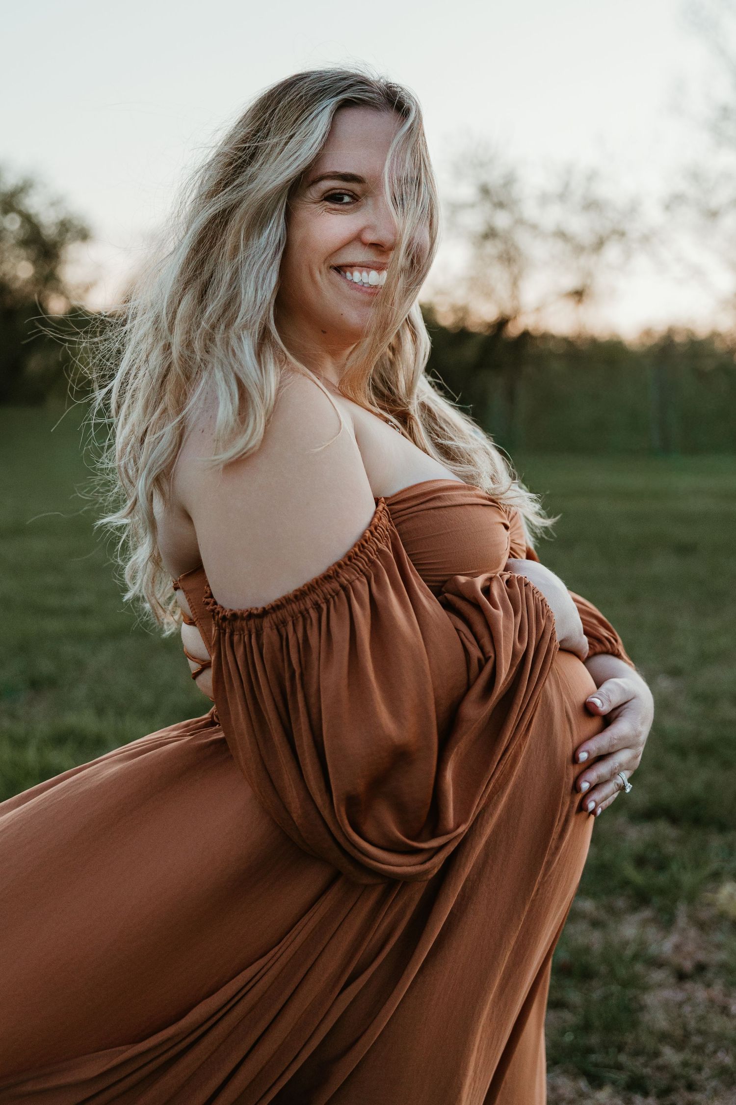 Smiling pregnant woman in rust off-shoulder gown poses outdoors at golden hour, cradling her baby bump.