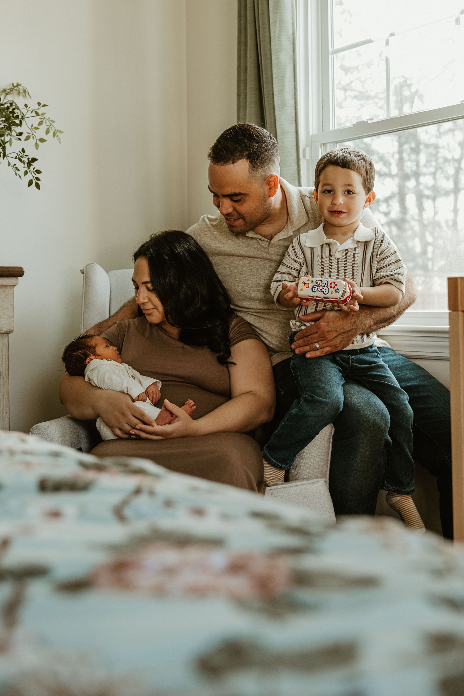 Family of four sitting by window, parents admiring newborn baby while toddler son holds a toy box.