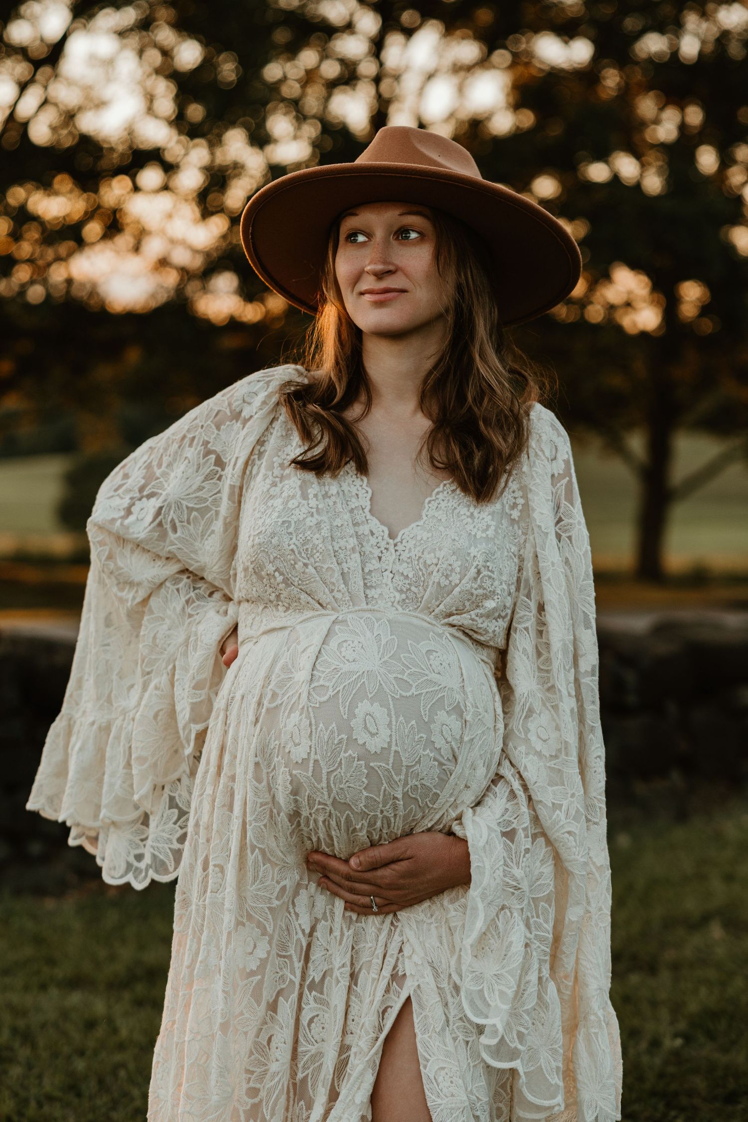 Maternity portrait featuring a brown hat and detailed white lace dress in golden sunlight.