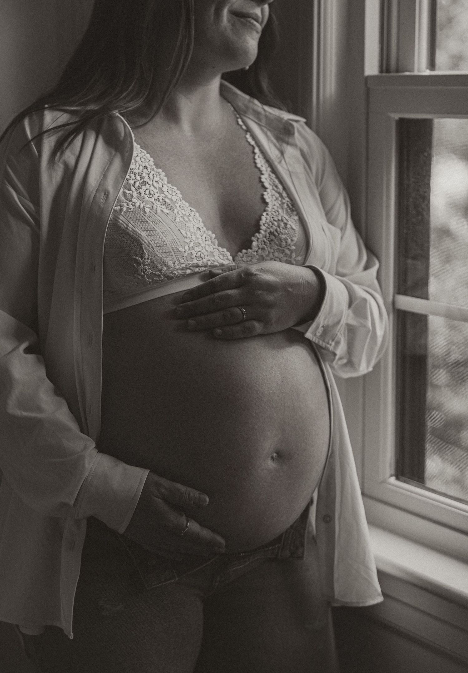 Black and white maternity photo of pregnant woman in lace bralette and open shirt cradling baby bump near window.