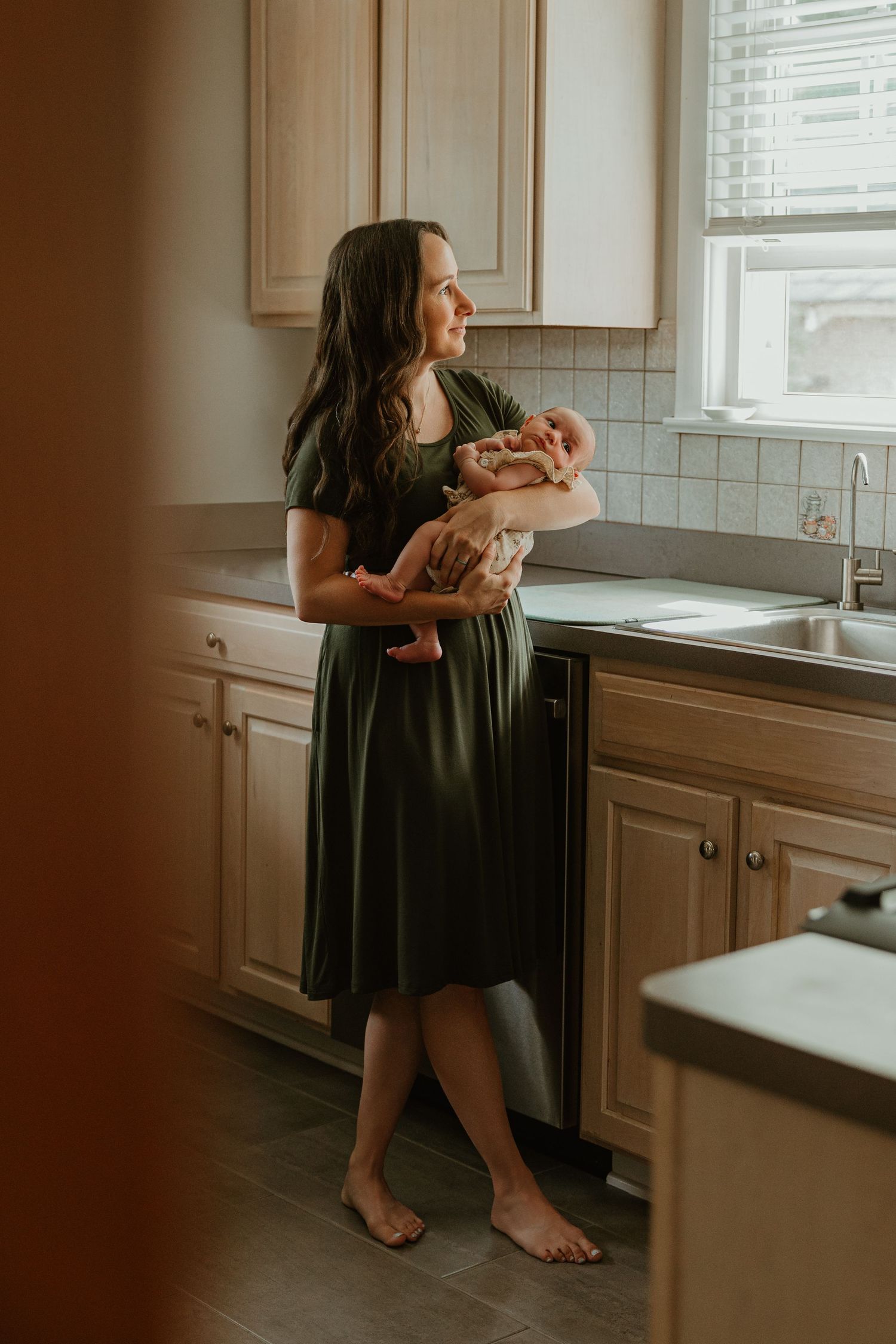 A barefoot mother in an olive green dress holds her newborn while gazing out a kitchen window.