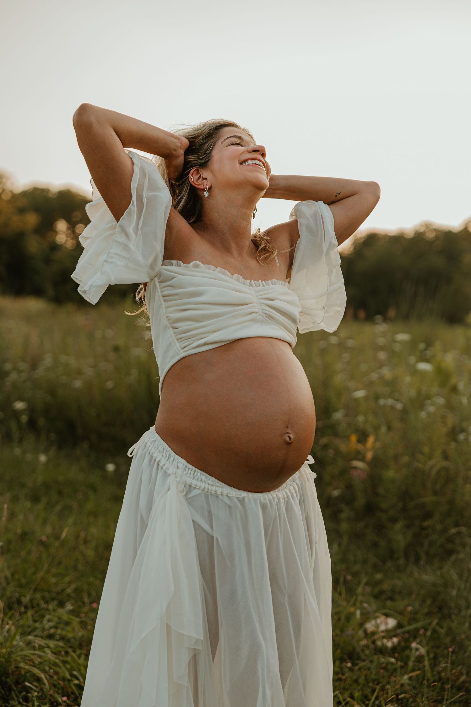 Smiling pregnant woman in white crop top and skirt stands in sunlit meadow, hands behind head, showcasing baby bump.