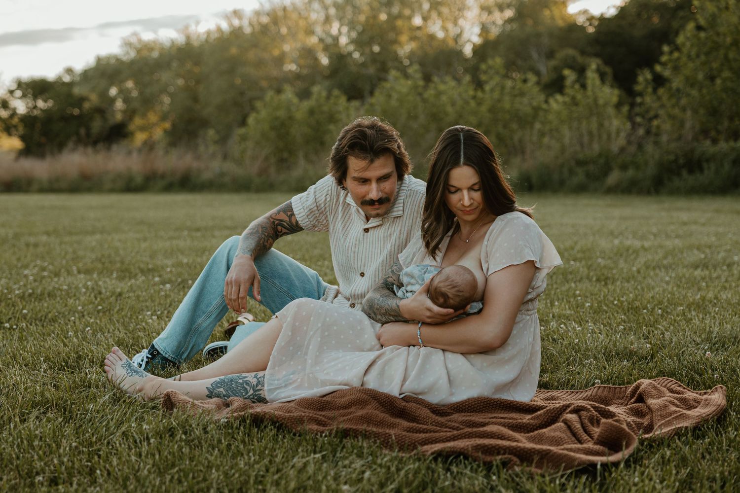 A couple sits on a blanket in a green field, cradling their newborn baby during a golden hour outdoor photoshoot.