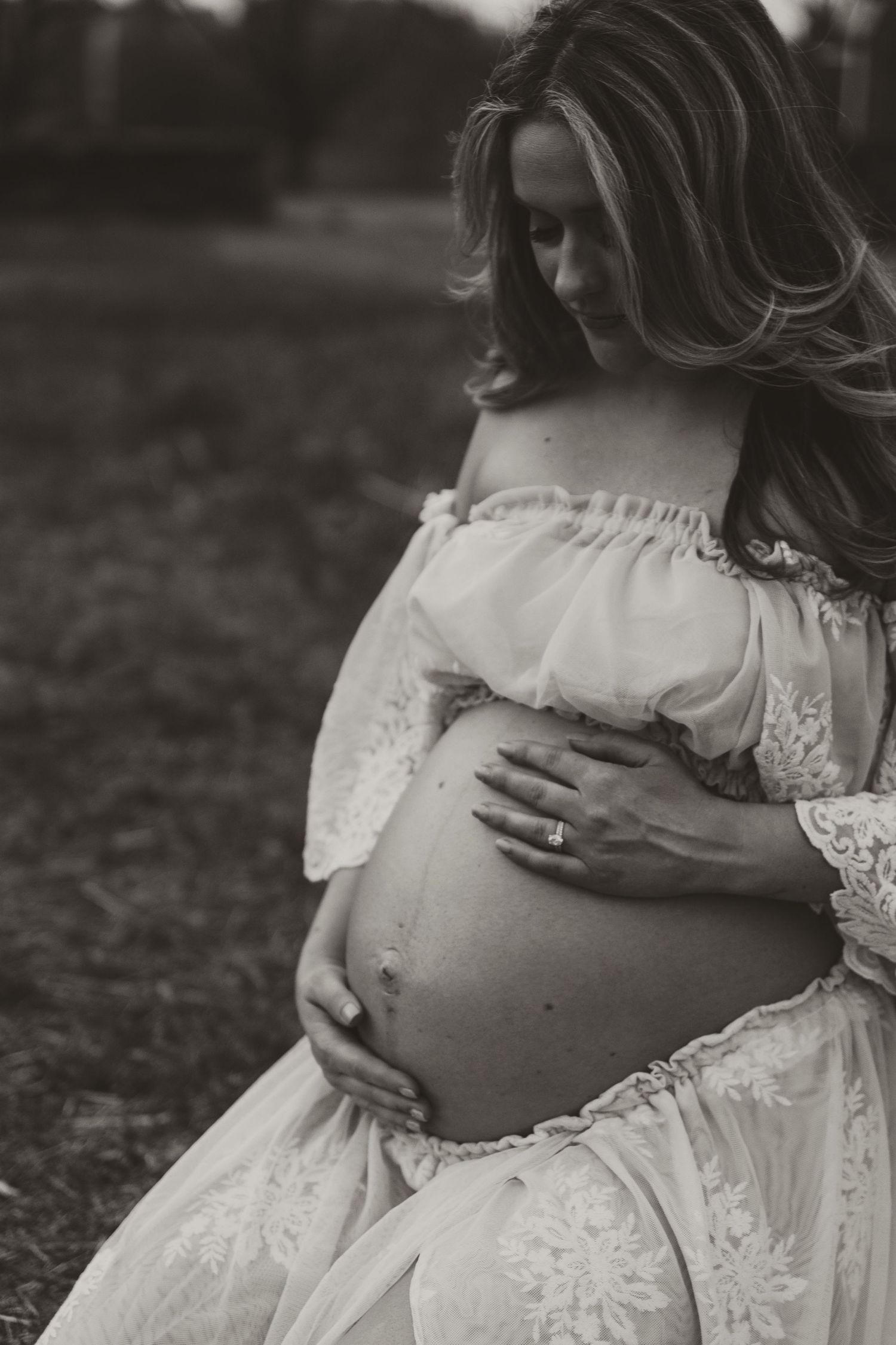 Artistic black and white maternity photo of expectant mother in lace dress cradling her belly in outdoor setting.