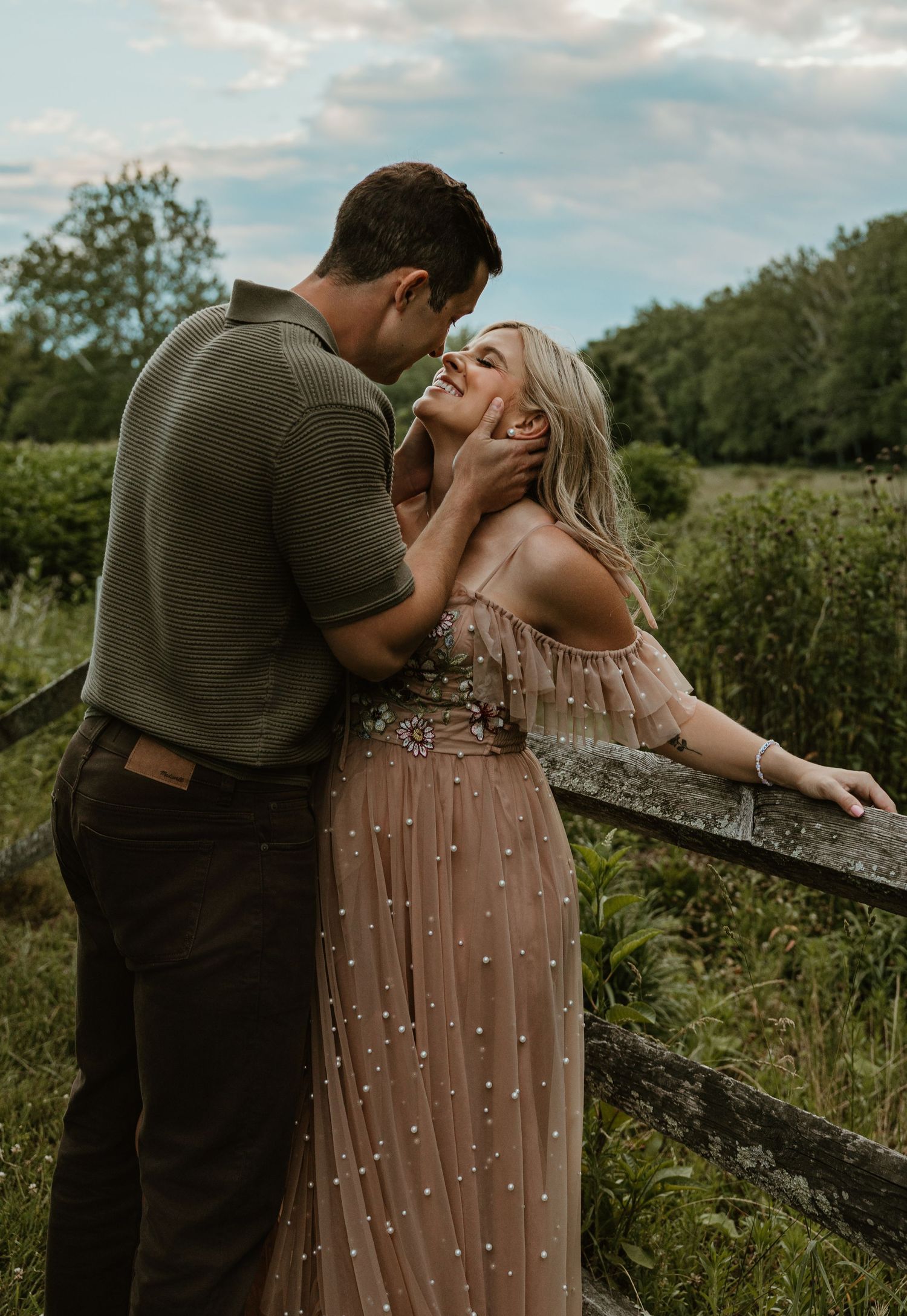 Romantic couple embracing by a rustic fence in a lush green field, woman in pearl-embellished blush gown smiling up at man.