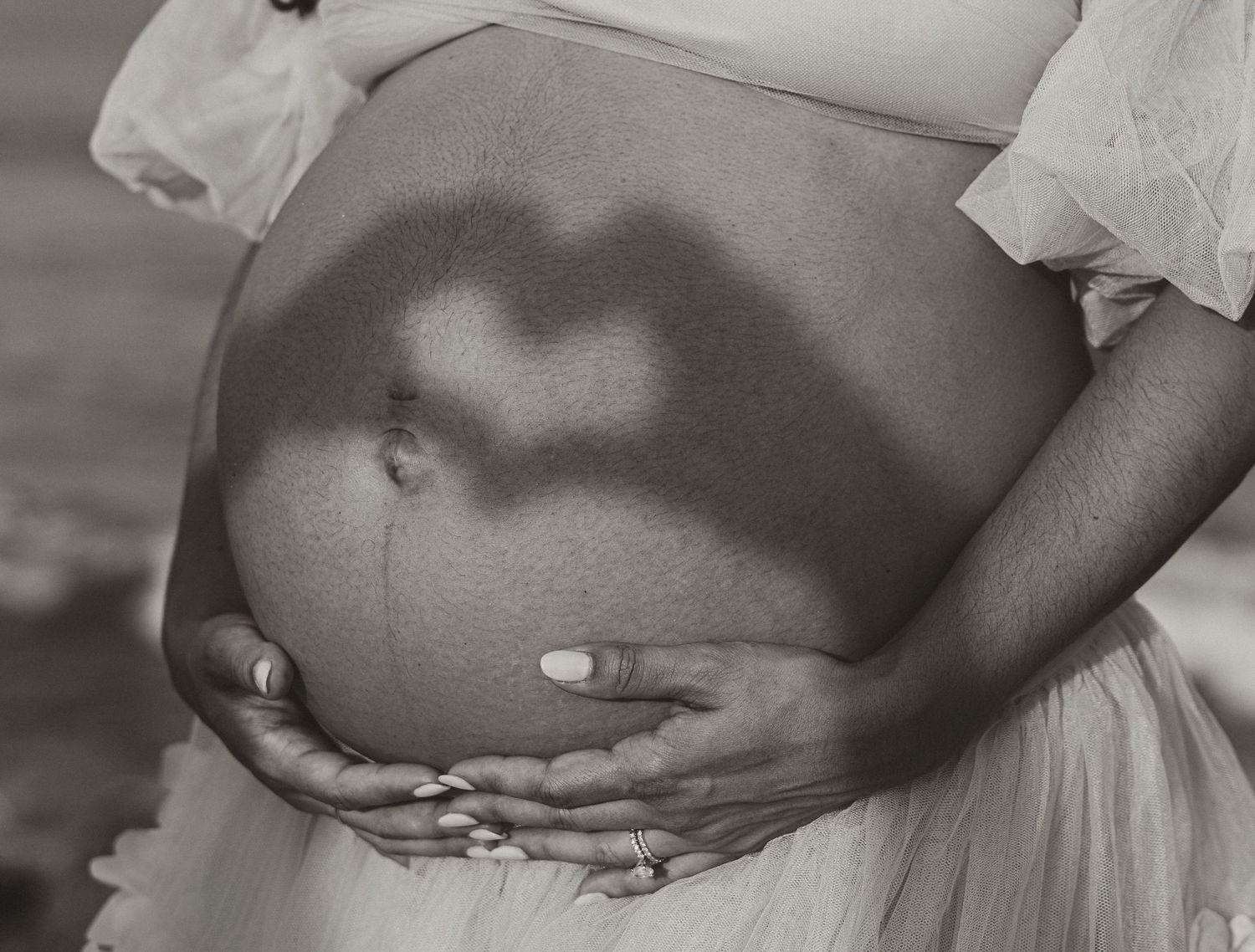Black and white photo of pregnant woman's belly with heart-shaped shadow, cradling bump with ringed hands.