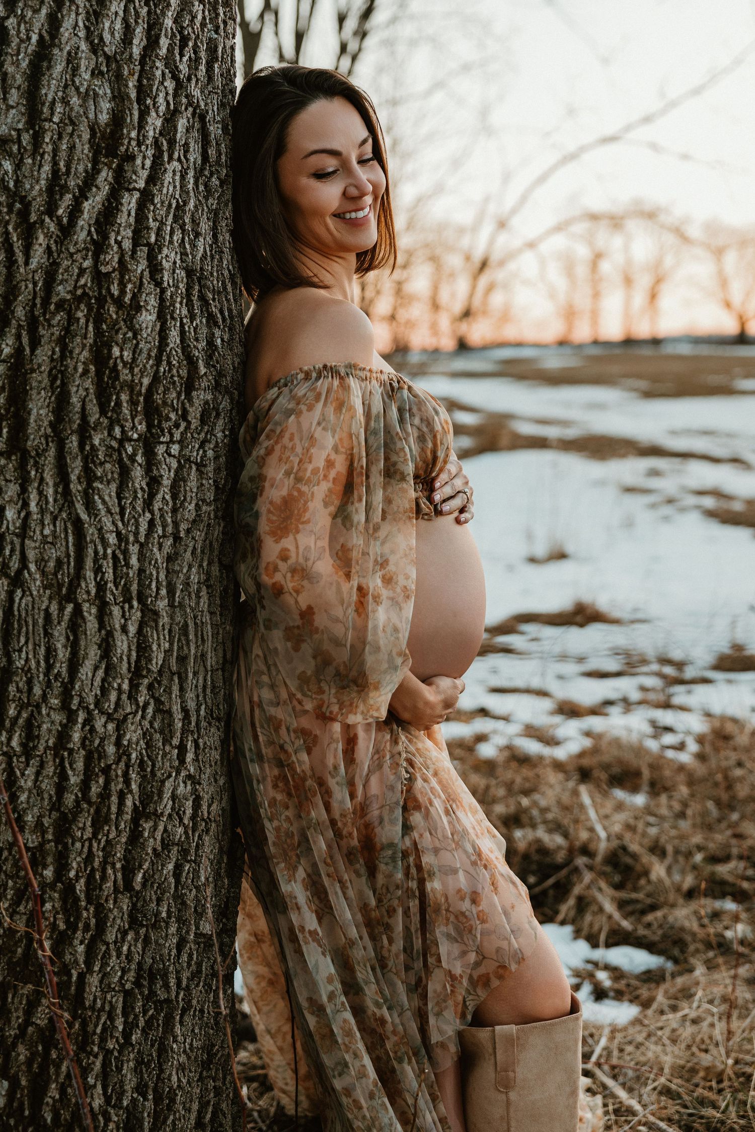 Maternity photoshoot in a snowy winter landscape featuring a floral dress and natural outdoor setting at sunset.