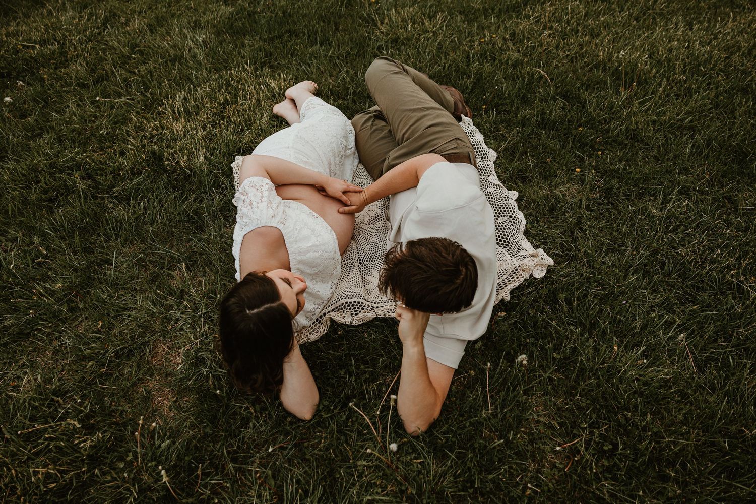 Aerial view of couple lying on lace blanket in green grass, holding hands in romantic outdoor photo session.