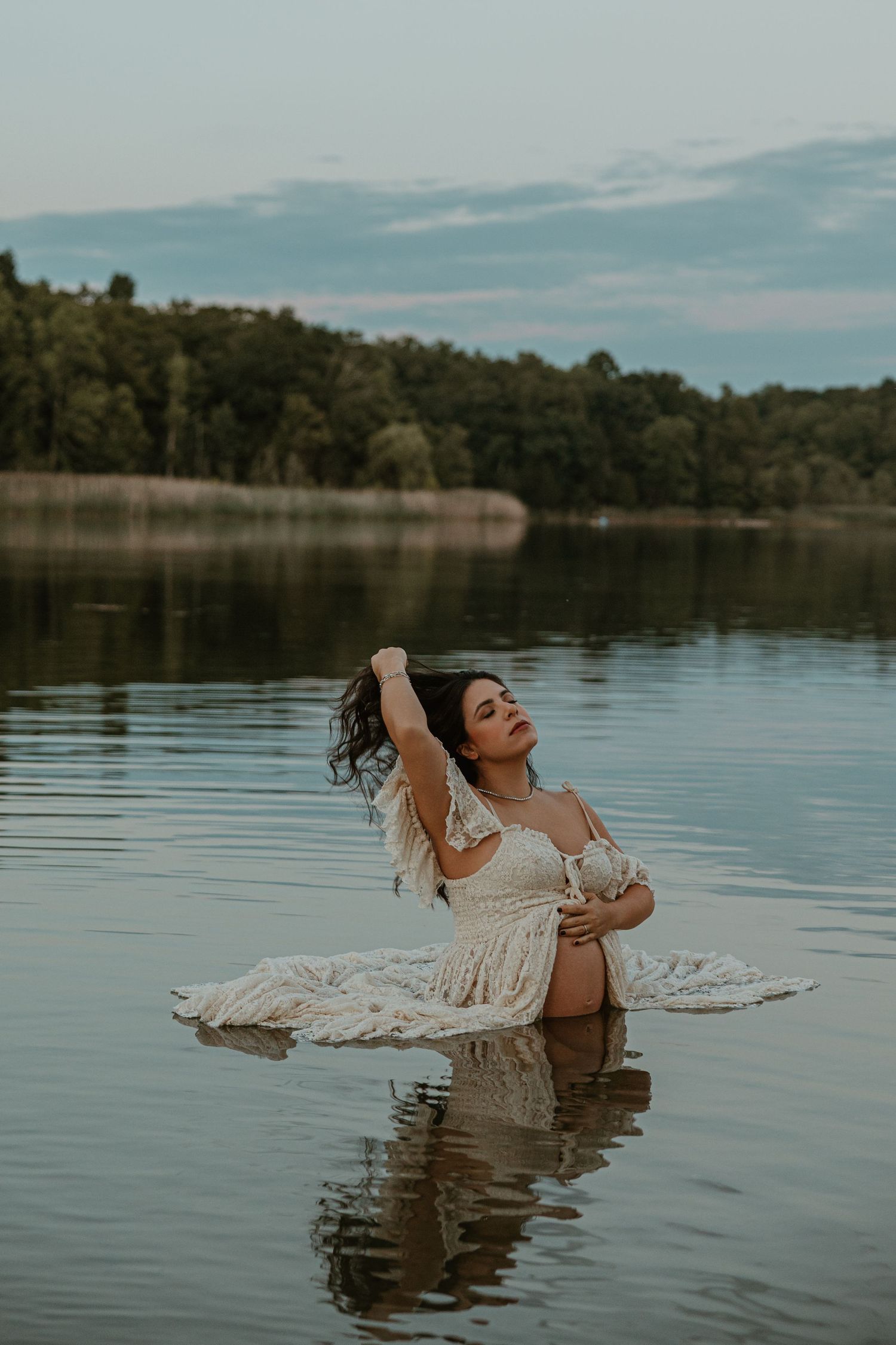 Pregnant woman in white lace dress sitting in calm lake water, surrounded by lush green forest, in a serene maternity photo.