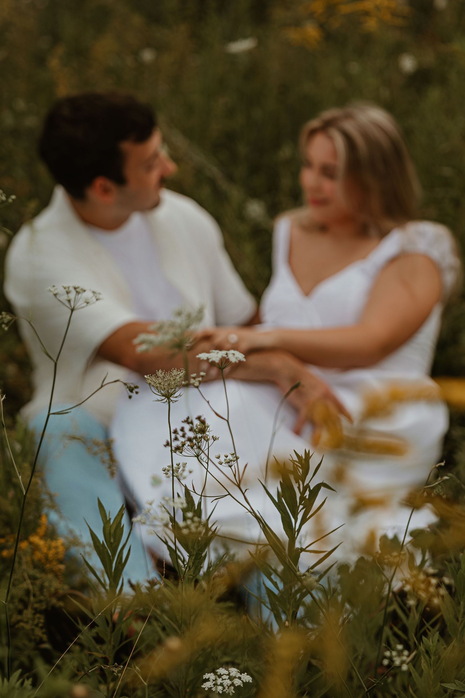Couple in white sitting together in a wildflower meadow, holding hands, with soft bokeh foreground flowers.
